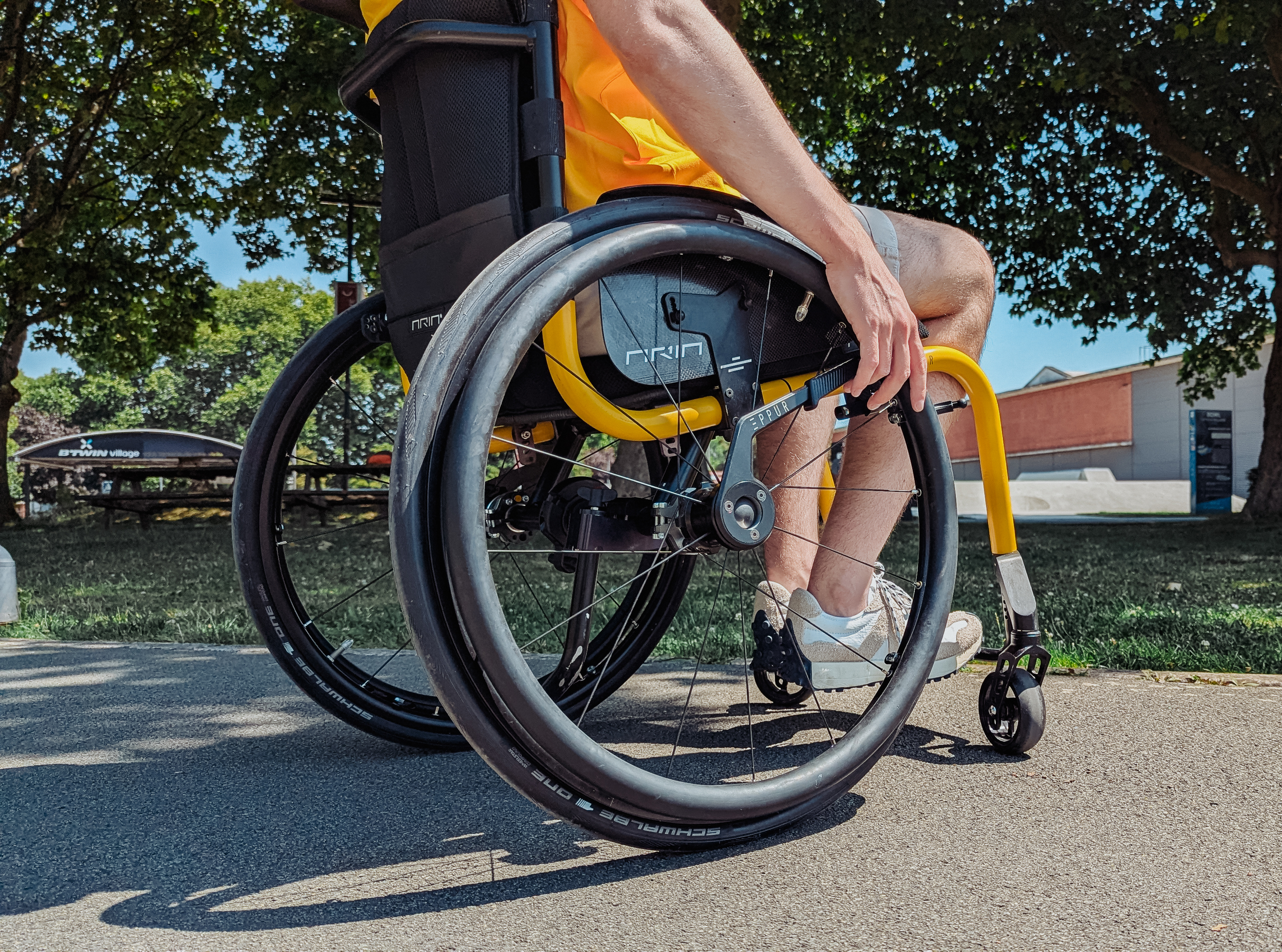 Close-up of a person seated in a yellow wheelchair outdoors, holding the wheel with one hand. The person is wearing a yellow shirt, shorts, and sneakers. The background features trees and a building.
