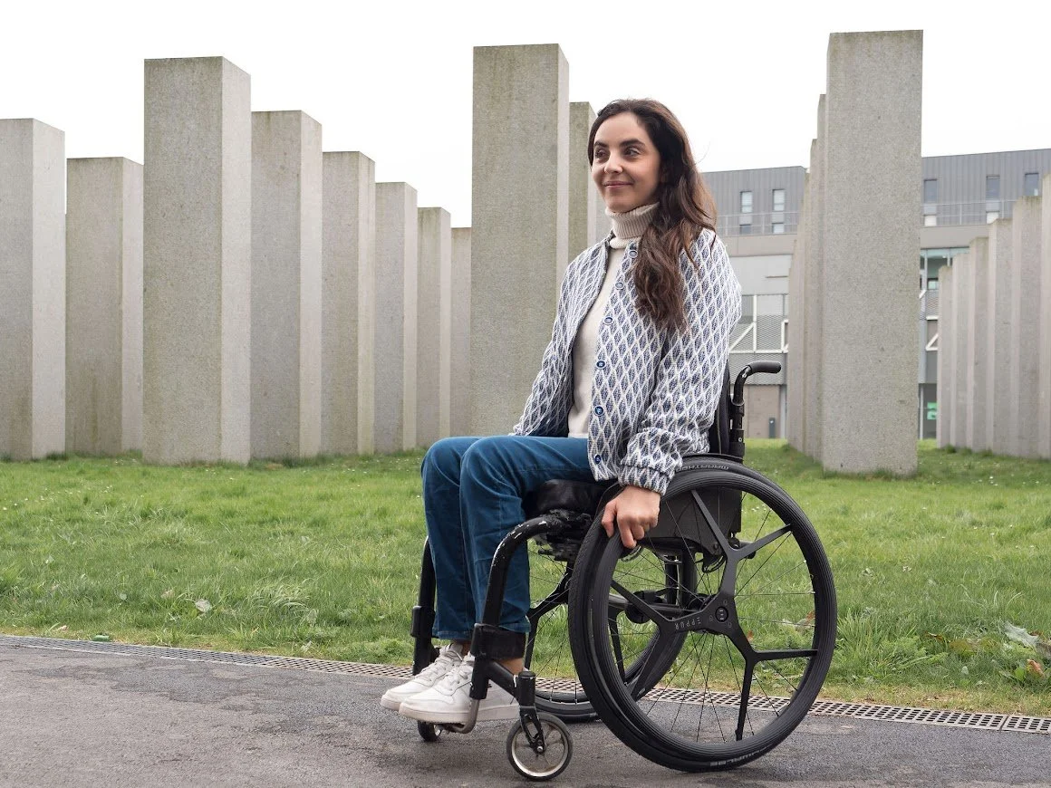 A woman in a wheelchair outdoors with concrete pillars and a modern building in the background.