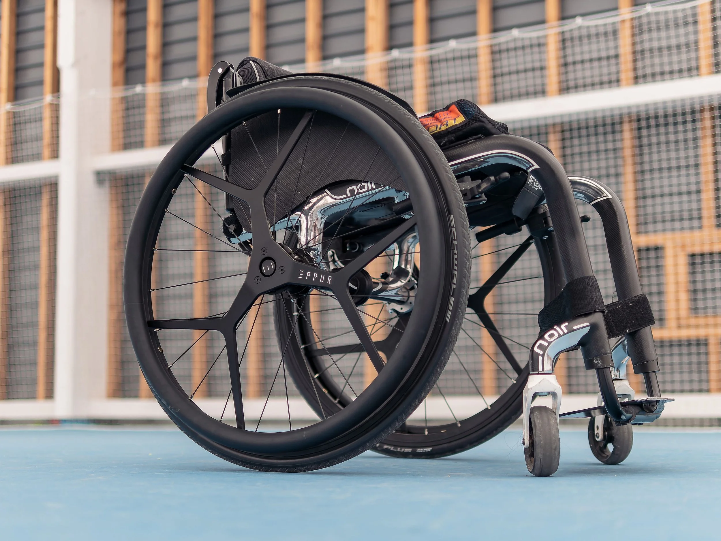 Wheelchair on a sports court with a metal fence and wooden structure in the background.