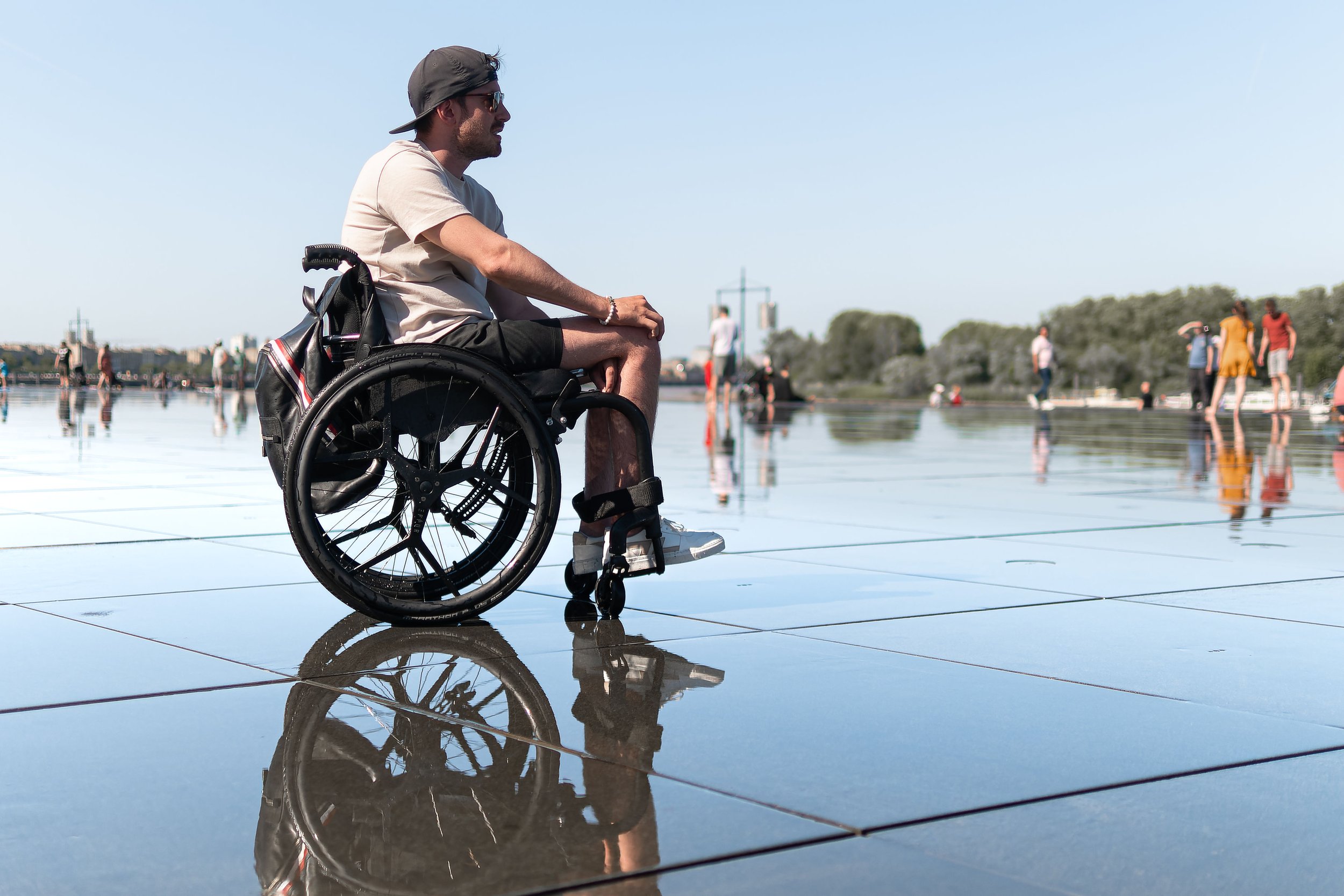 Man in a wheelchair sitting on a shiny, reflective surface outdoors with people walking in the background.