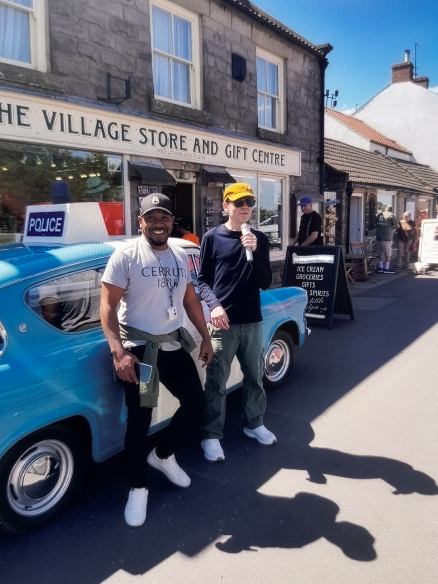 Two men standing outside a building in front of a vintage police car. One man is smiling and wearing a white shirt, baseball cap, and white sneakers. The other man is holding a microphone, wearing sunglasses, a yellow cap, and gray pants with white sneakers. The building behind them has a sign for the village store and gift center, with other people and shops visible in the background.