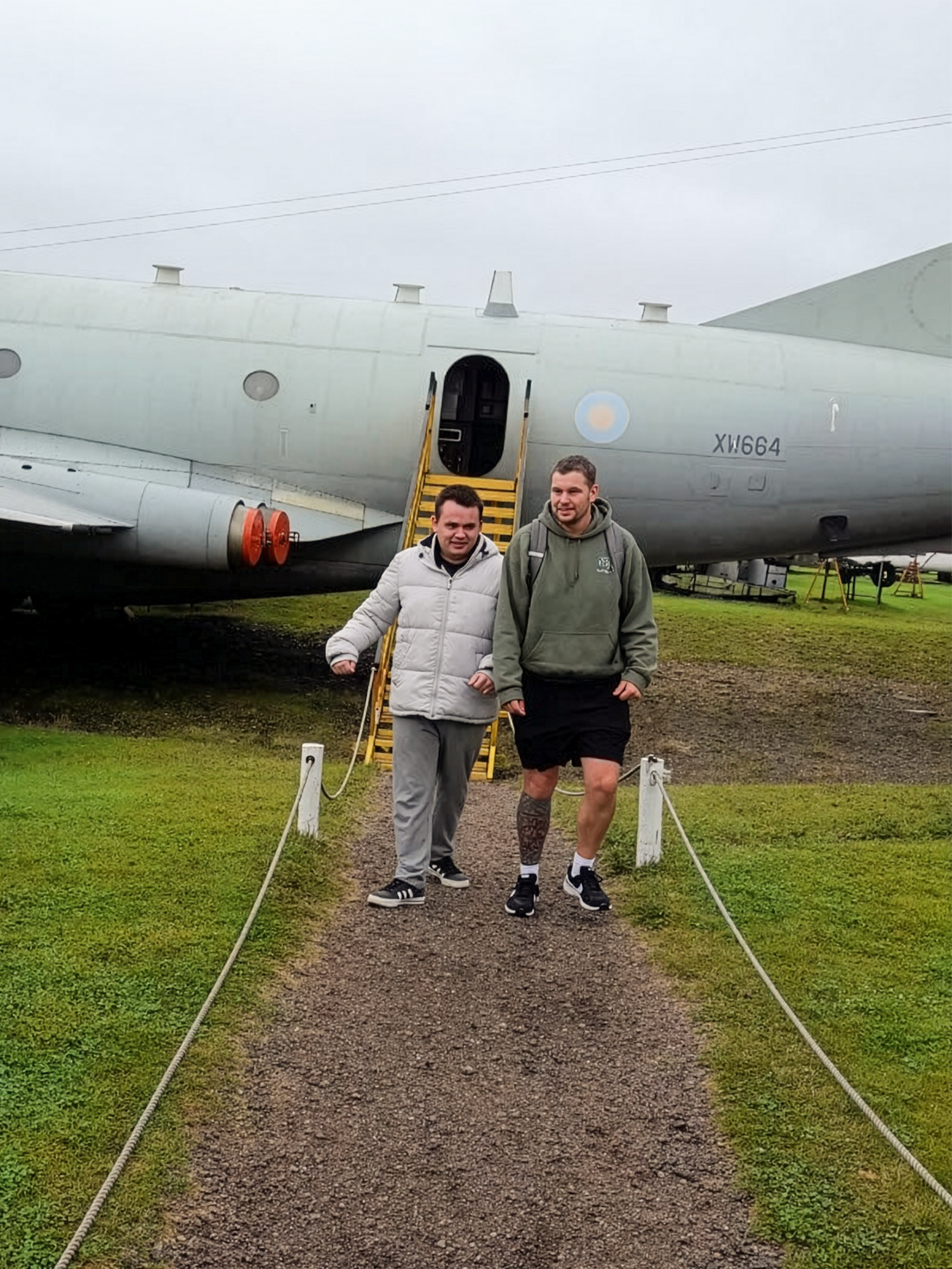 Two men walking away from a military aircraft, one wearing a hoodie and the other a jacket, on a gravel path with grass on either side.