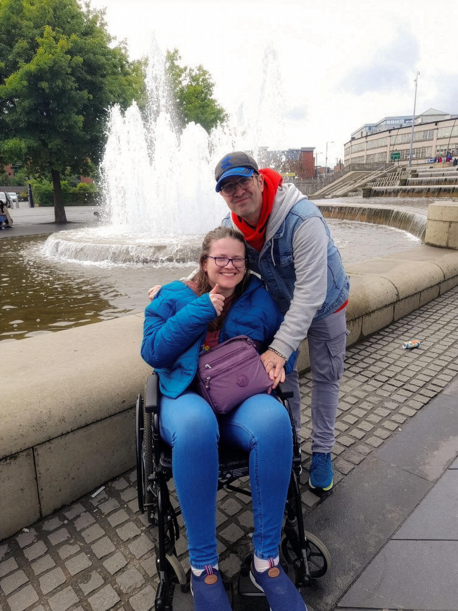 A man and a woman, with the woman using a wheelchair, posing together in front of a water fountain in an urban area. The man is wearing a denim vest, red hoodie, and a cap, while the woman is wearing glasses and a blue jacket. They are smiling at the camera.