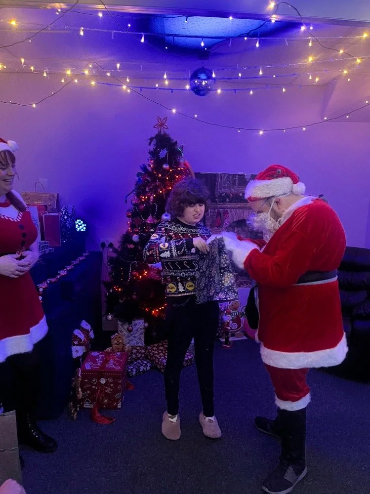 A young girl receives a gift from a person dressed as Santa Claus at a decorated Christmas party with a Christmas tree, presents, and holiday lights.