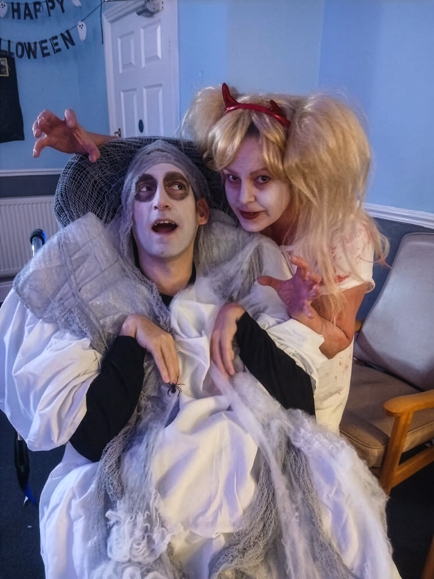 Two people dressed in Halloween costumes; one as a clown with messy blond hair and red devil horns, and the other as a zombie bride with gray hair and dark eye makeup, sitting together in a decorated room.