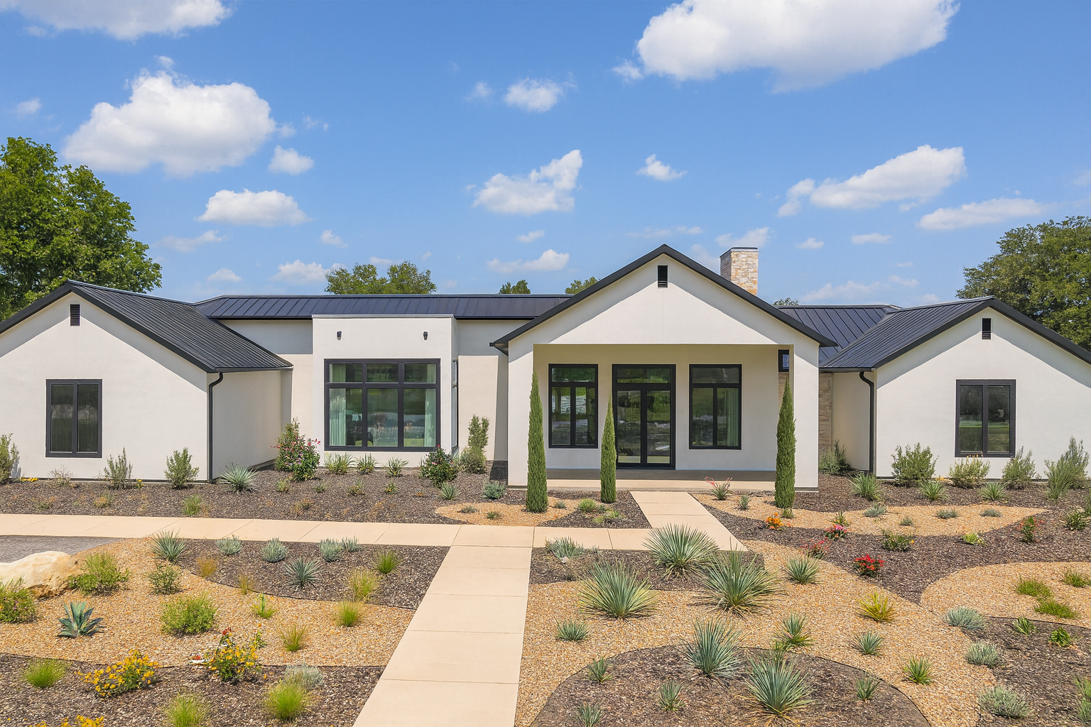 Modern single-story house with white exterior, black trim, and a black metal roof, surrounded by desert landscaping with drought-tolerant plants and a concrete walkway.