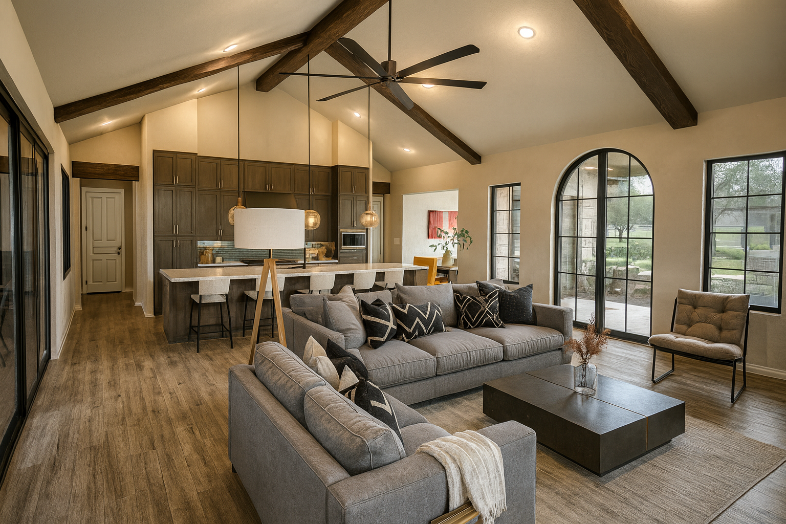 Open-concept living room with gray sofas, black and beige pillows, a wooden coffee table with a vase, wood flooring, large arched windows, and a kitchen with dark cabinets, a bar with beige chairs, pendant lighting, and a ceiling fan with dark blades