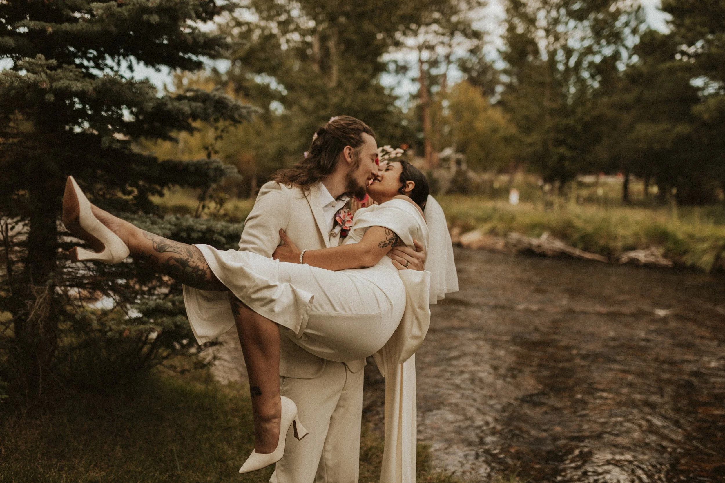 A couple in wedding attire, with the man in a cream suit and the woman in a white dress, kissing near a river surrounded by trees. The woman is being carried by the man and is wearing white high heels.
