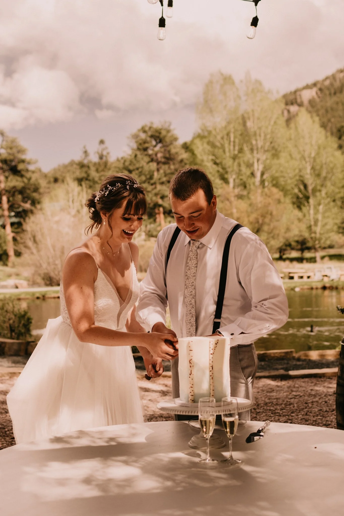 A bride and groom cutting a wedding cake outdoors near a pond with trees in the background, under hanging string lights.