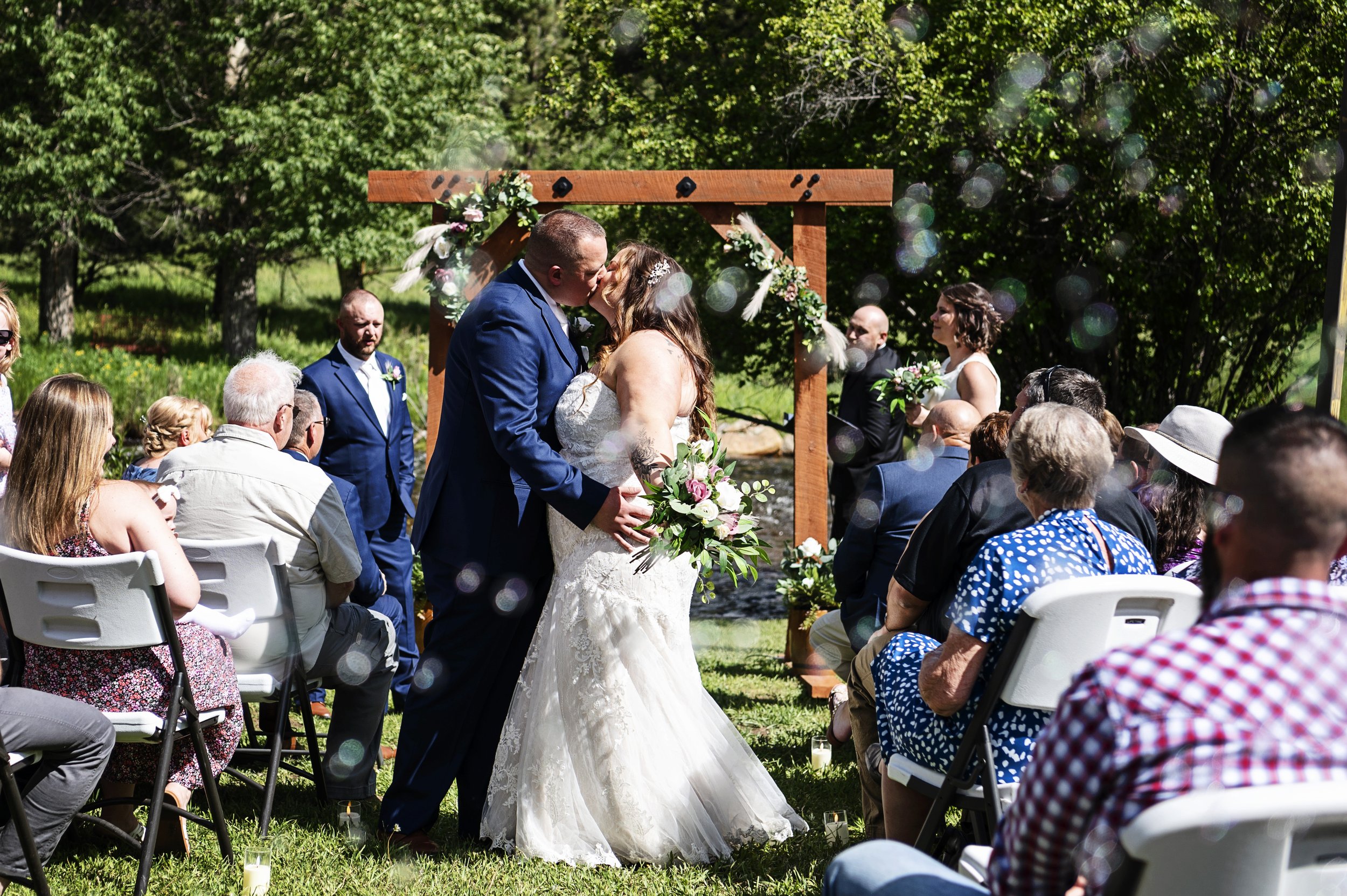 A couple sharing a kiss during an outdoor wedding ceremony with guests seated around them, a wooden arch decorated with flowers, trees in the background, and sunlight creating bokeh effects.