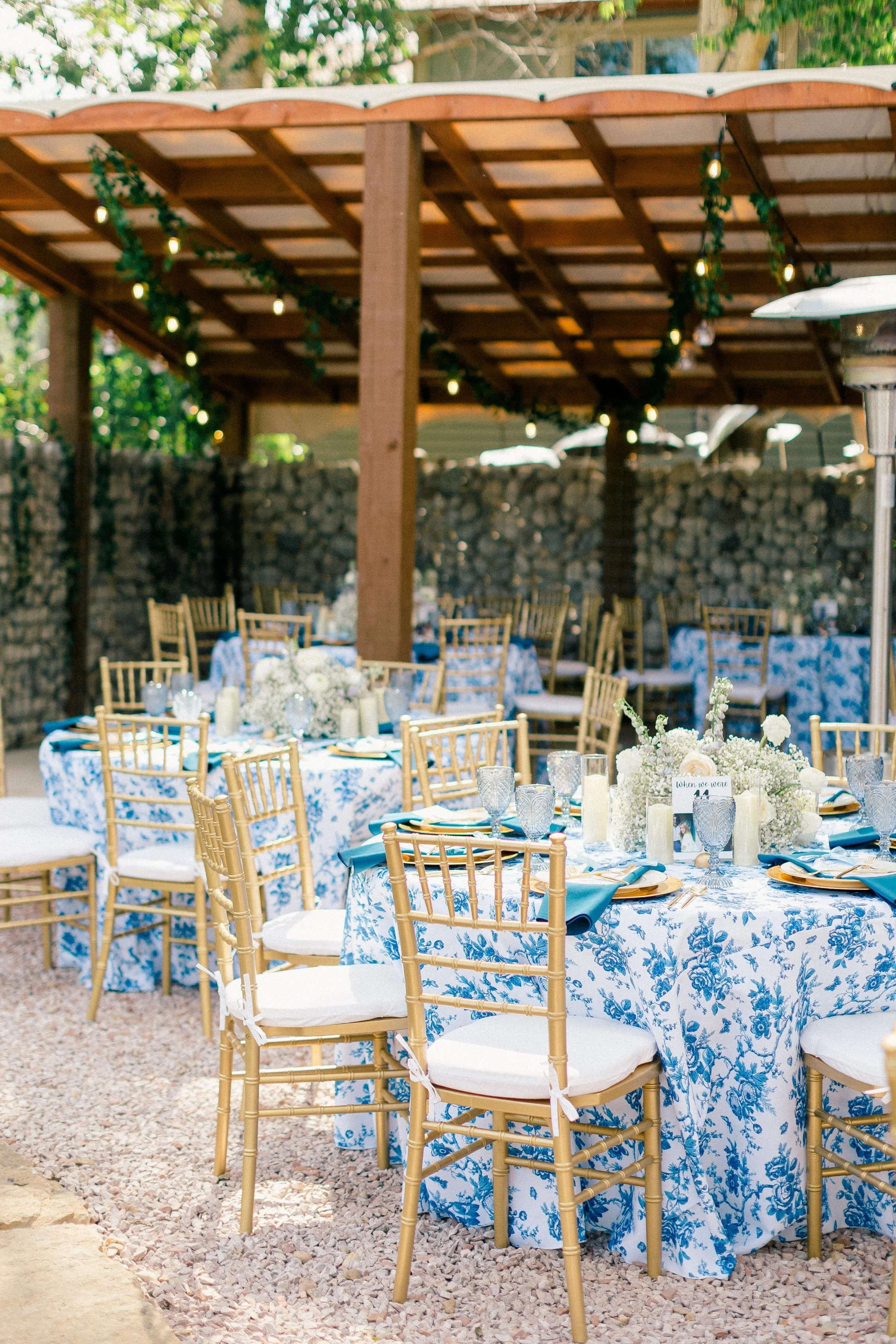 Outdoor wedding or event setup with round tables covered in blue and white floral tablecloths, gold chairs with white cushions, floral centerpieces, and string lights under a wooden pergola surrounded by a stone wall.