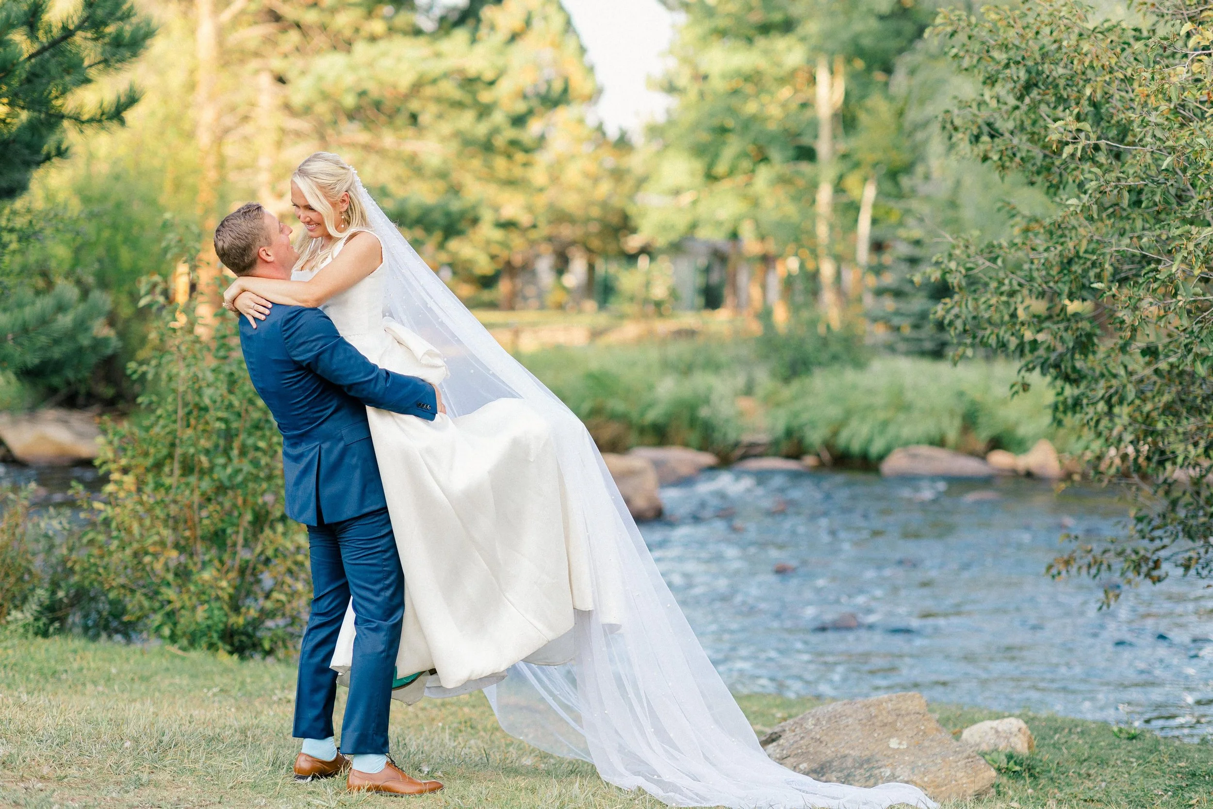 A groom lifting a bride by a creek in a lush outdoor setting during daytime, surrounded by trees and greenery.