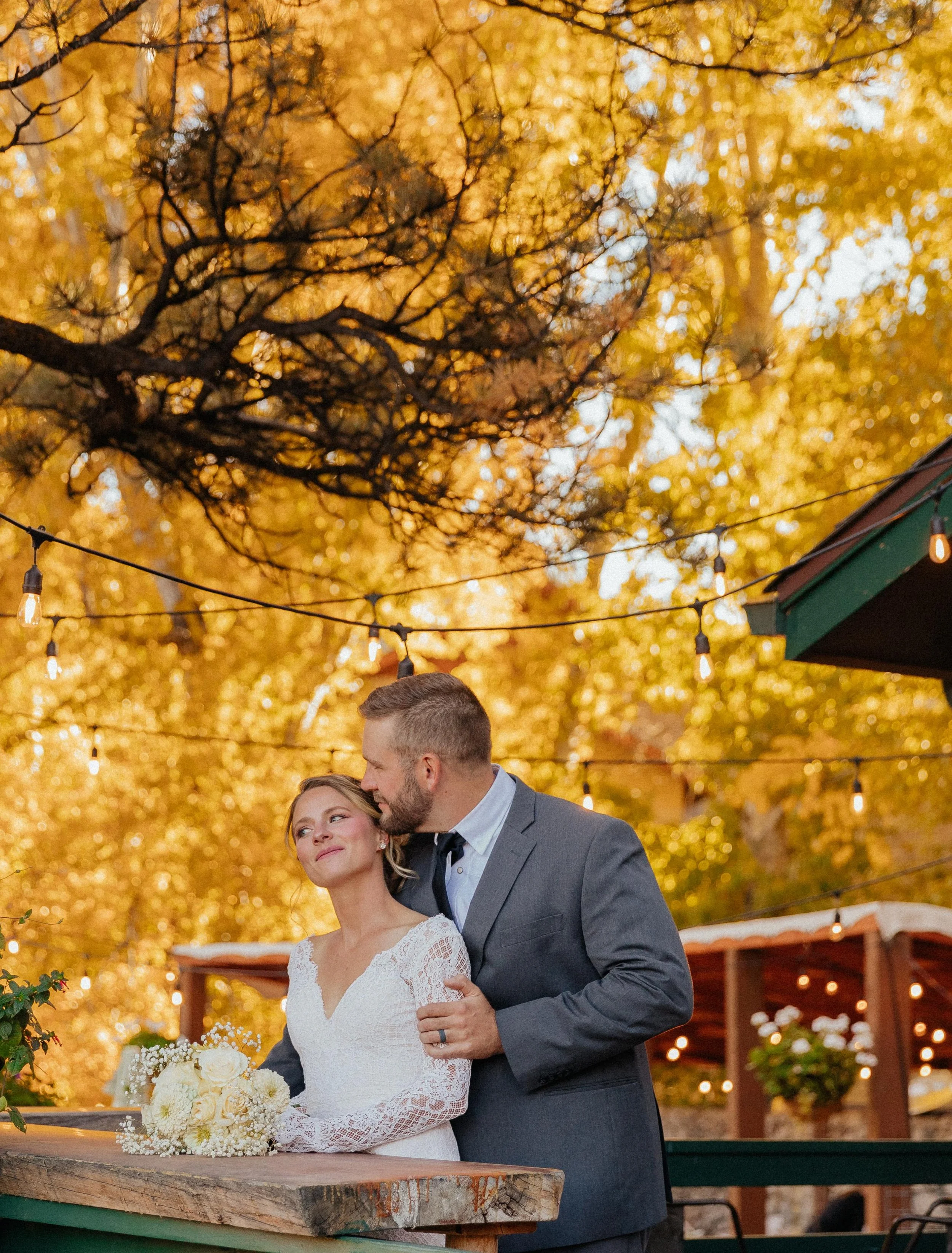 A bride and groom at an outdoor wedding reception with autumn leaves and string lights overhead. The bride is holding a bouquet of white flowers, and the groom is leaning in close to her.