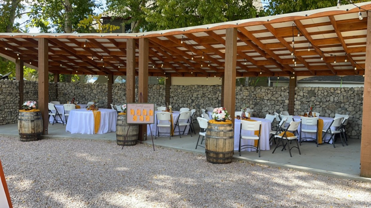 Outdoor wedding reception area with round tables covered in white tablecloths and yellow table runners, decorated with flower arrangements in vases. Wooden barrels are used as table stands, and a sign with table number options is present. The area is partially enclosed with a wooden roof and string lights, with a stone wall background and gravel ground.