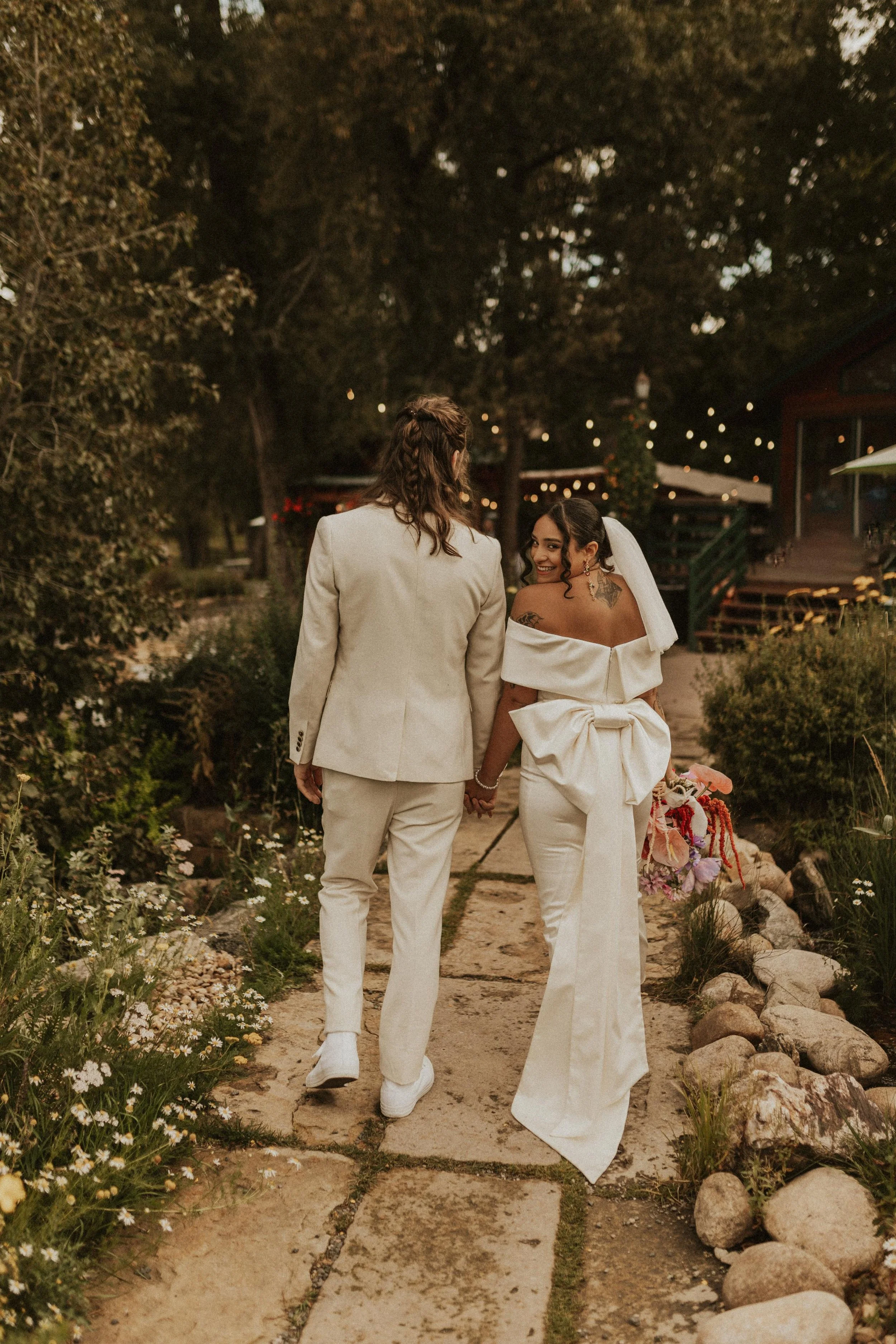 A bride and groom holding hands and walking down a garden pathway during sunset, with string lights overhead, surrounded by greenery and flowers.