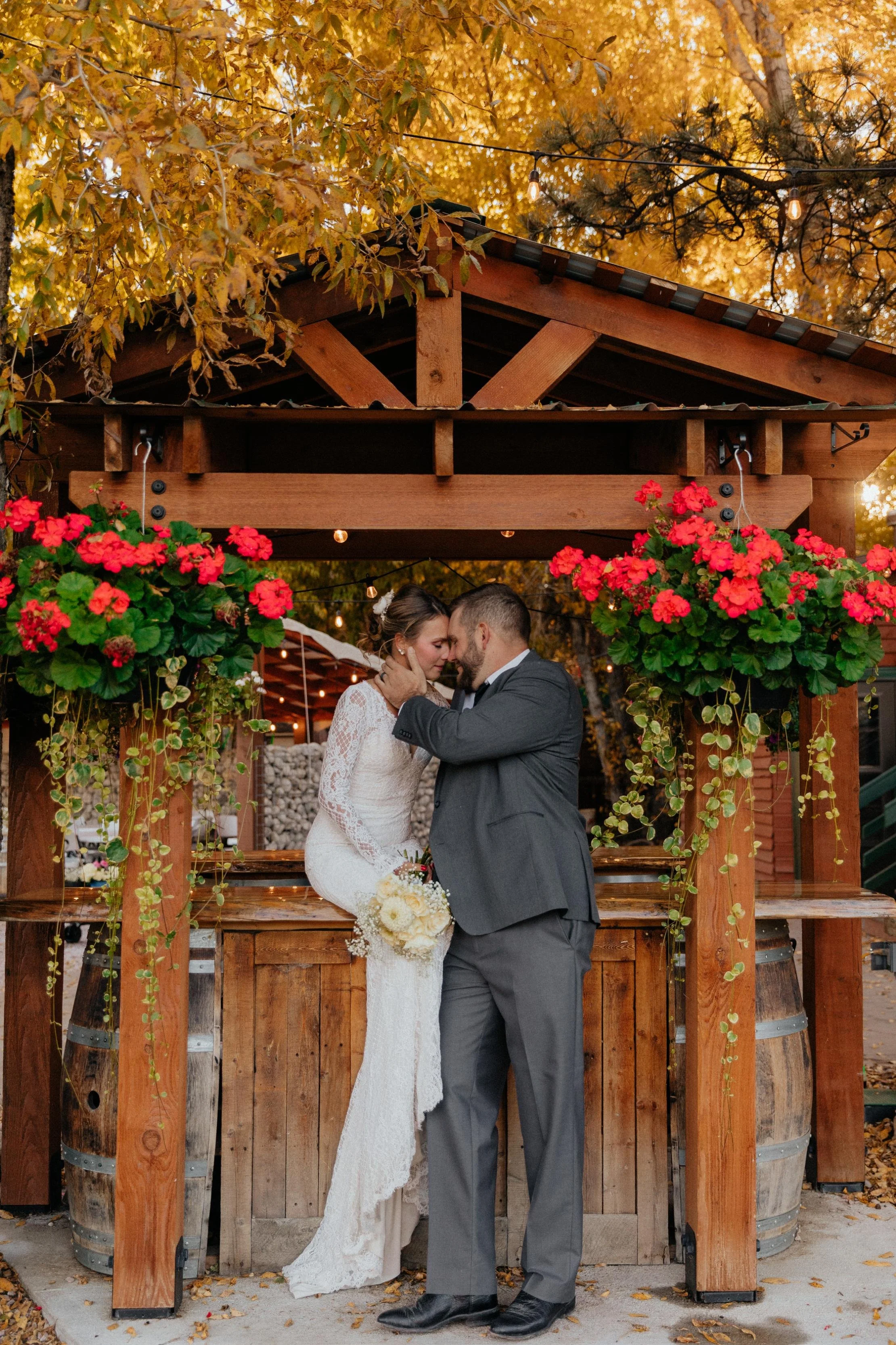A bride and groom sharing a kiss at a rustic outdoor wedding ceremony, with trees and string lights in the background and vibrant red flowers hanging on either side.