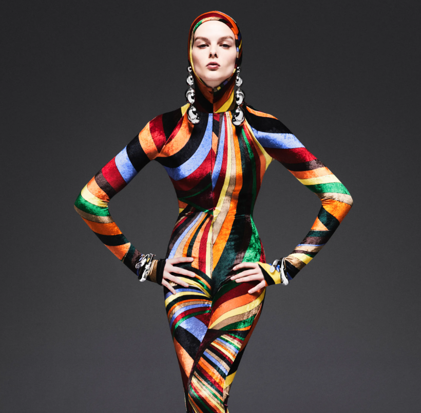 Une femme portant un vêtement intégré coloré avec des motifs de rayures dans plusieurs couleurs, accompagnée de boucles d'oreilles et bracelets assortis, posant avec les mains sur les hanches en face d'un fond gris foncé.