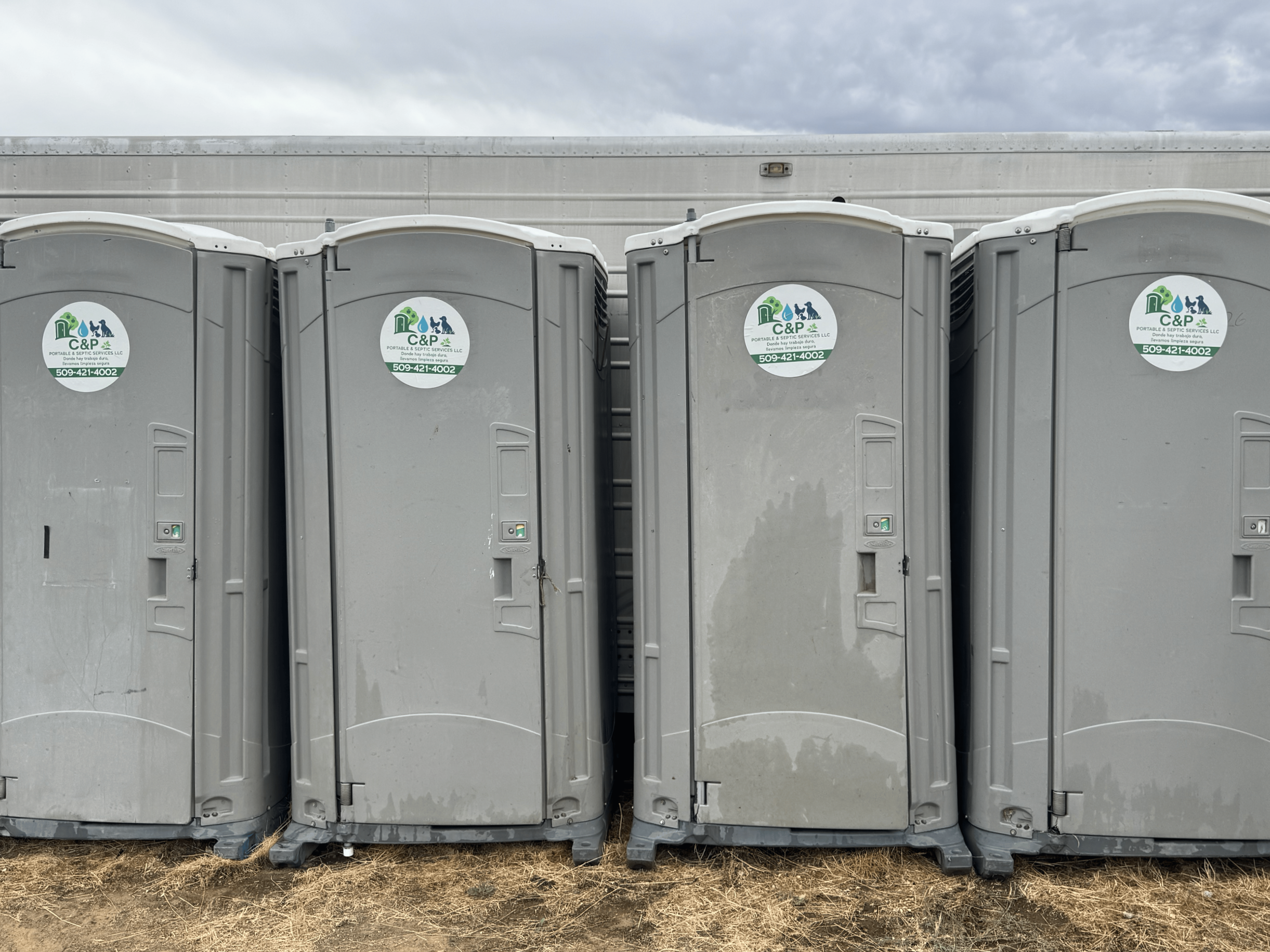 Four portable toilets with company stickers on each, positioned outdoors on a dirt ground with a cloudy sky in the background.