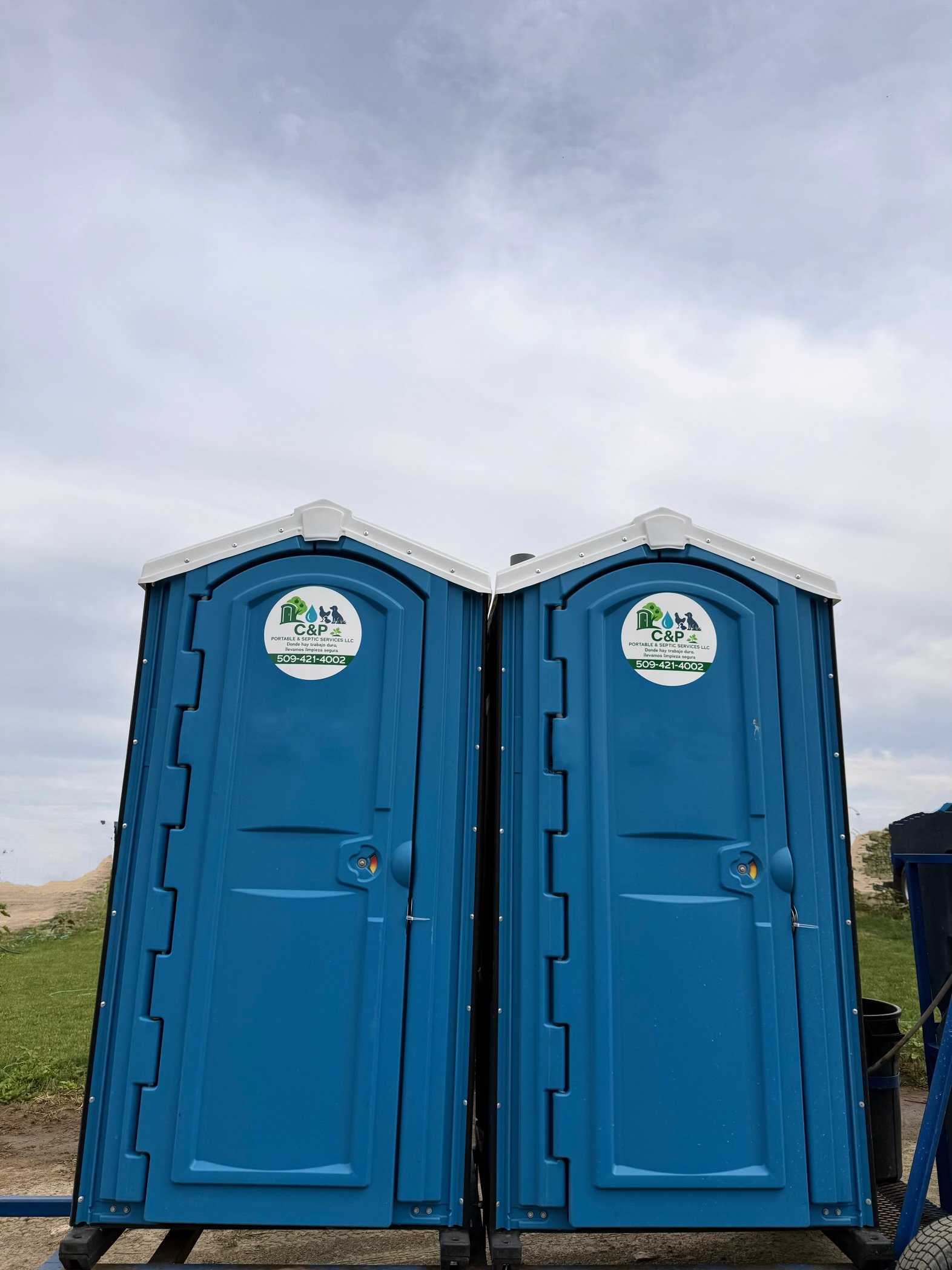 Two blue portable toilets with company logo and contact information, placed outdoors on grassy ground, under a cloudy sky.