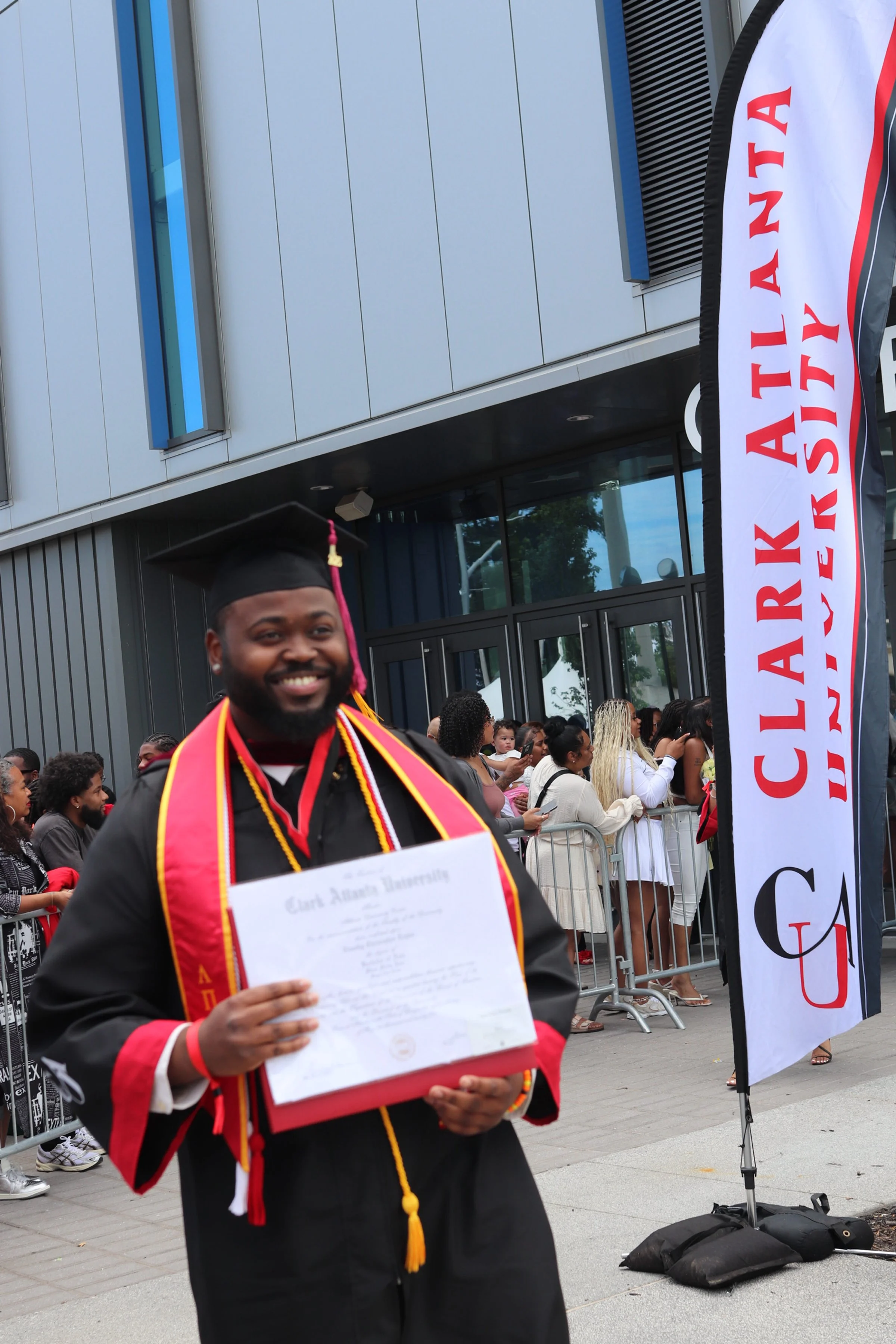 A man in graduation attire holding a diploma at a graduation ceremony outside a building, with a crowd of people and a banner in the background.