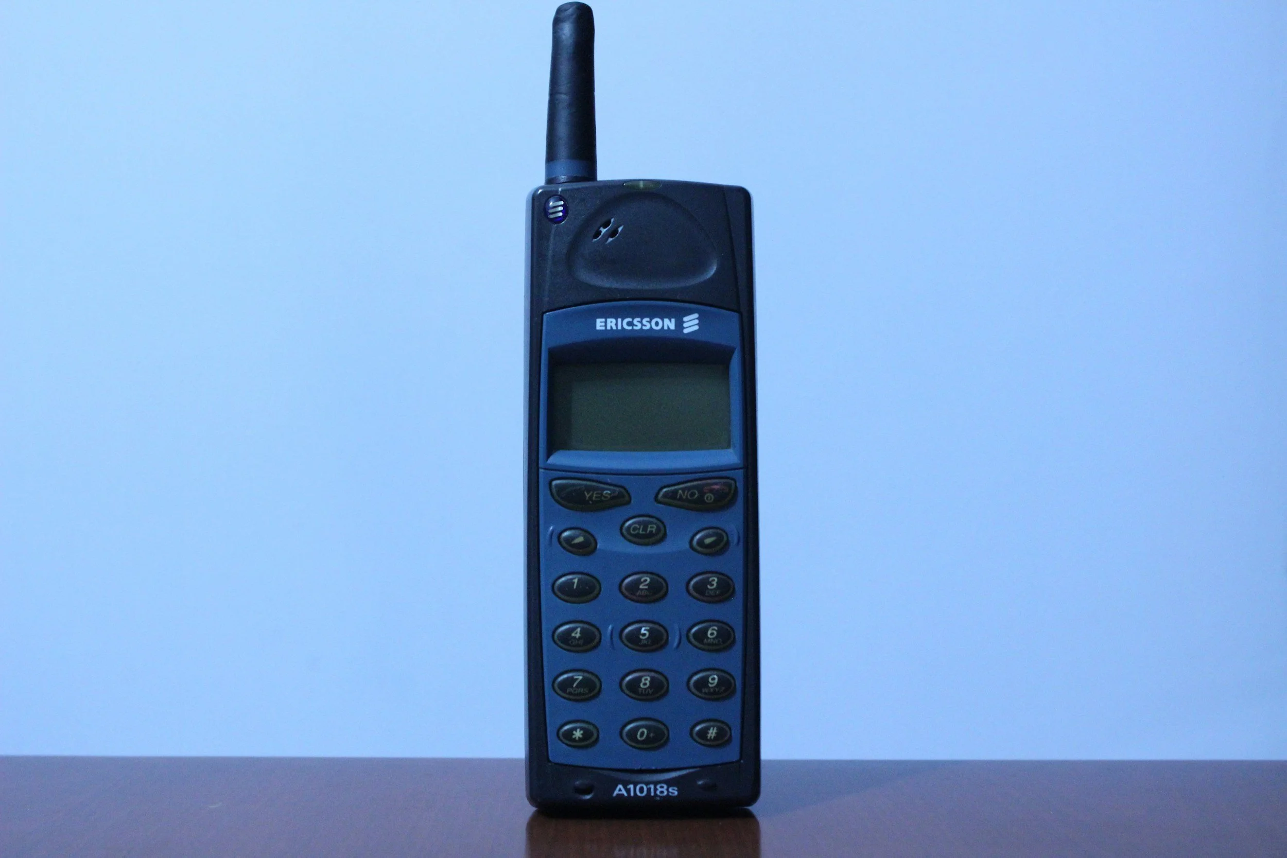 A black handheld mobile phone with an antenna, resting on a wooden surface. The phone has several buttons and a screen, with the brand name 'Ericsson' displayed on the upper part of the device.