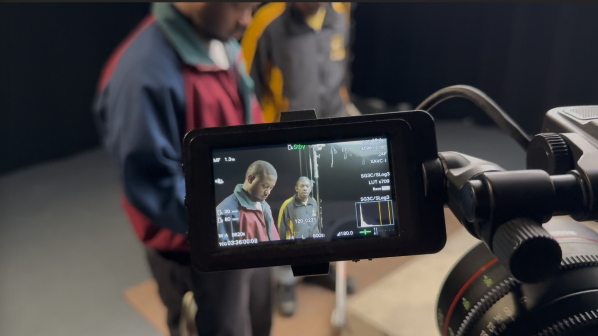 Close-up of a professional video camera recording two young men, one with a colorful jacket and the other in a gray and yellow jacket, in an indoor setting.