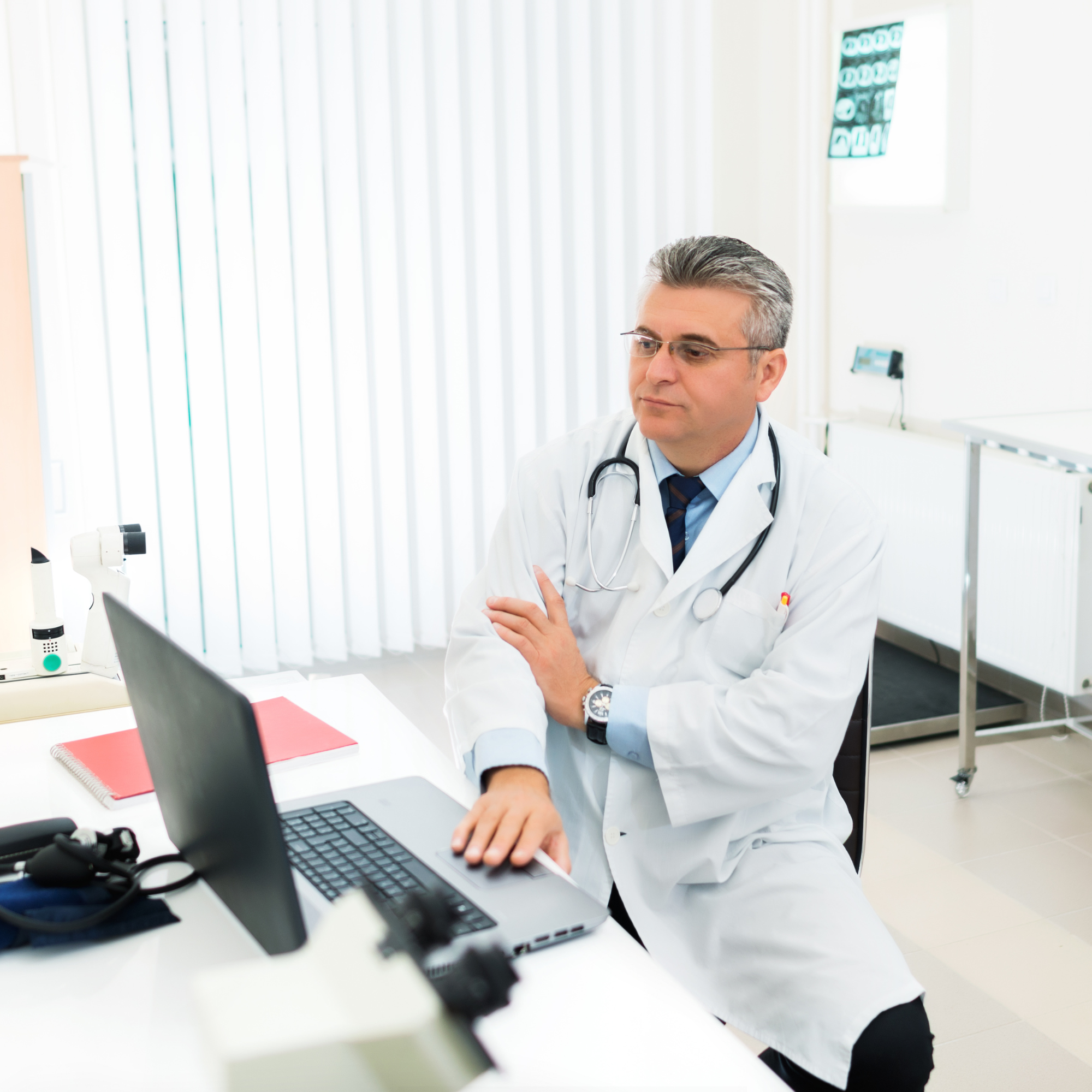 A male doctor in a white coat sitting at a desk with a laptop in a medical office, with a stethoscope around his neck and arms crossed, looking at the screen.