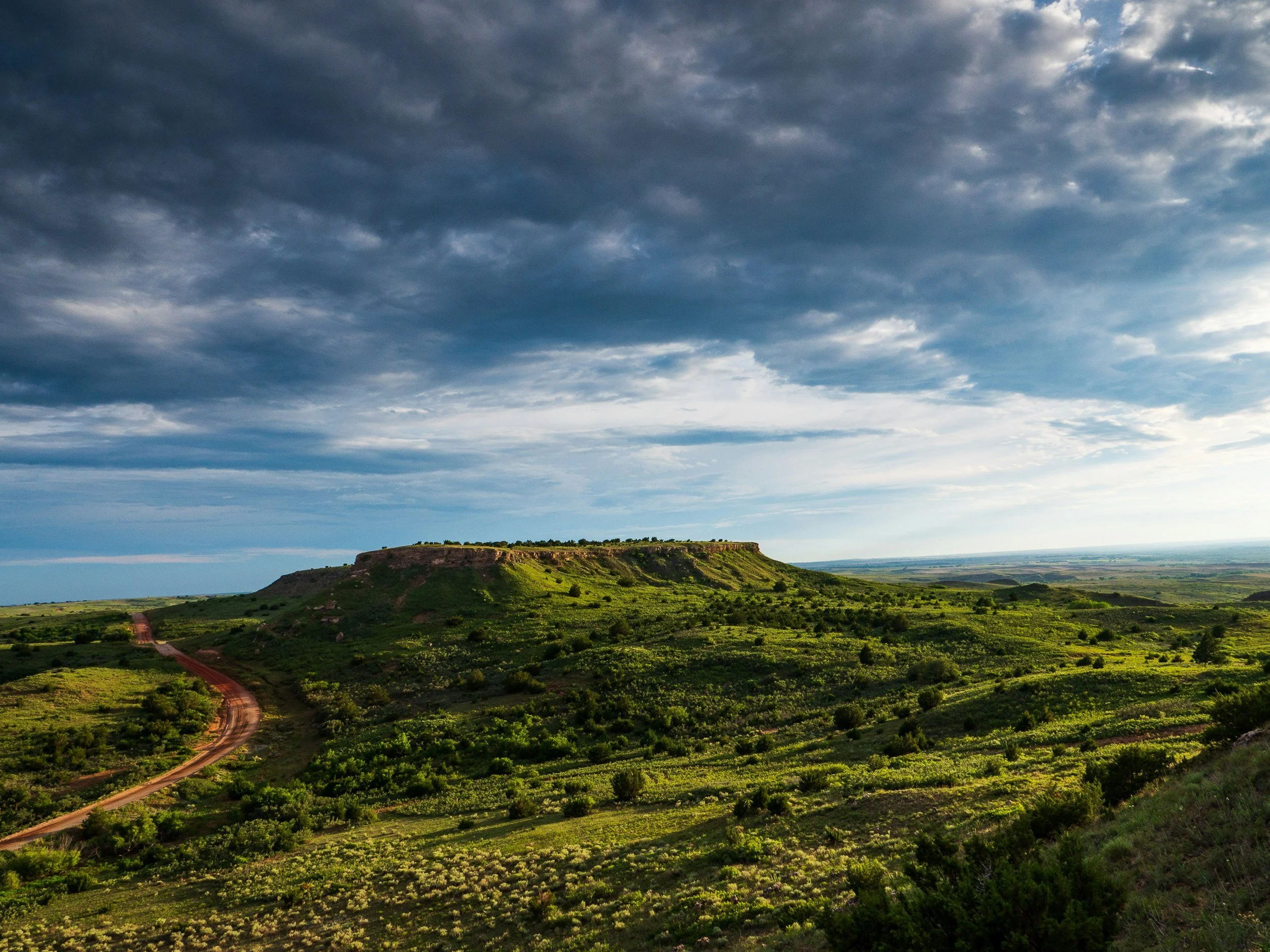 A scenic landscape featuring a flat-topped hill or mesa surrounded by green vegetation and a dirt road winding through the landscape, under a cloudy sky.
