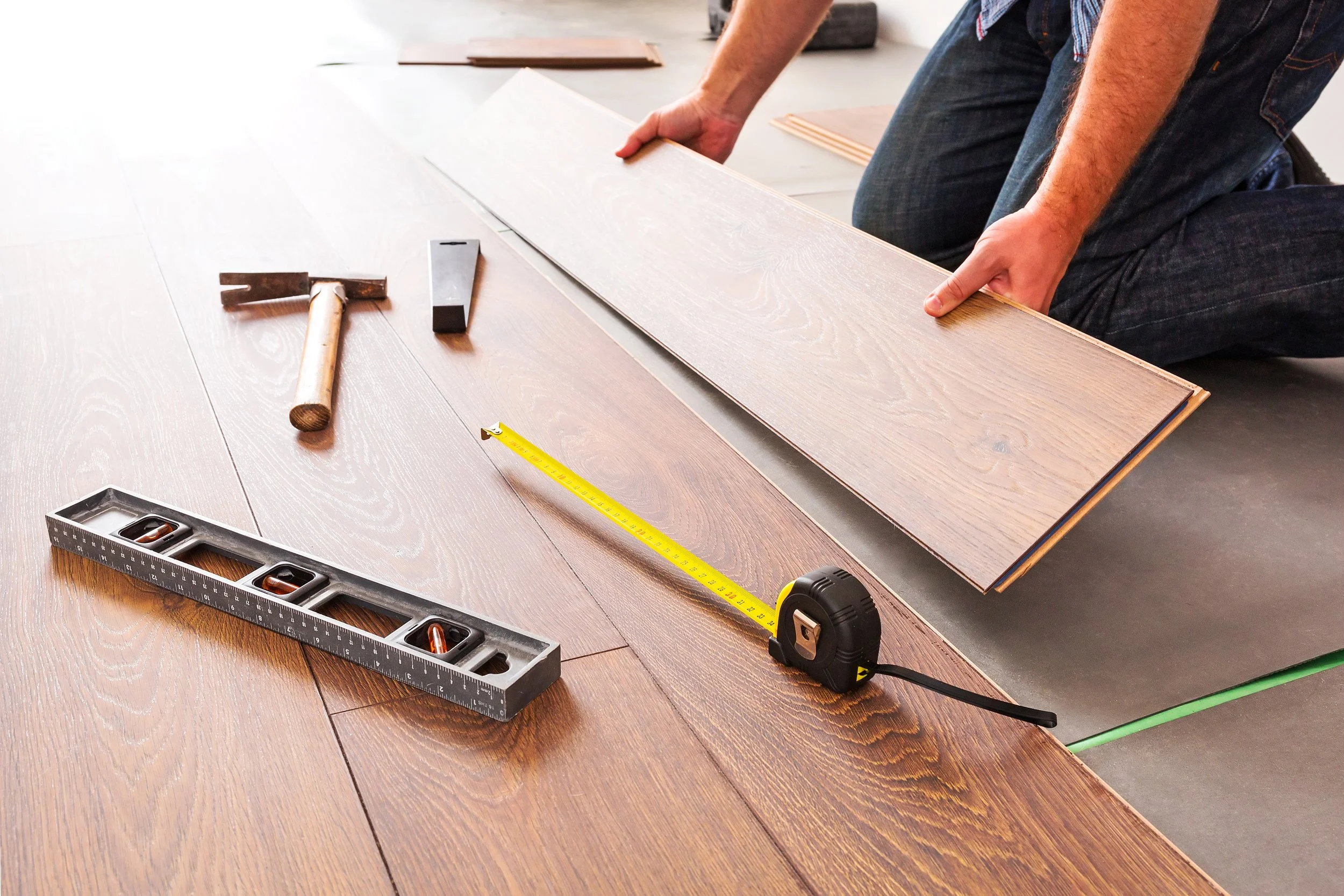 Person installing new wood flooring with tools including a hammer, measuring tape, level, and a piece of wood.