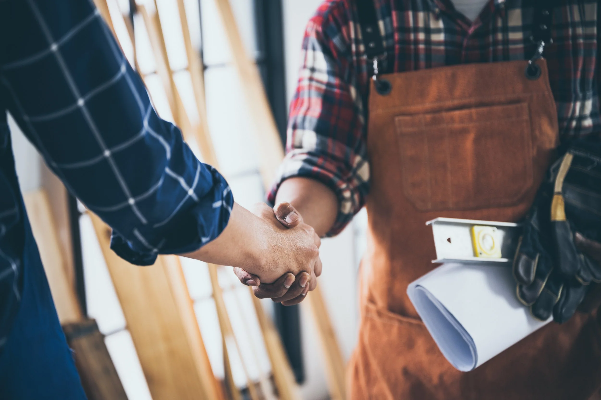 Two people shaking hands, one person is wearing a plaid shirt and apron, holding a rolled-up document and a level tool.