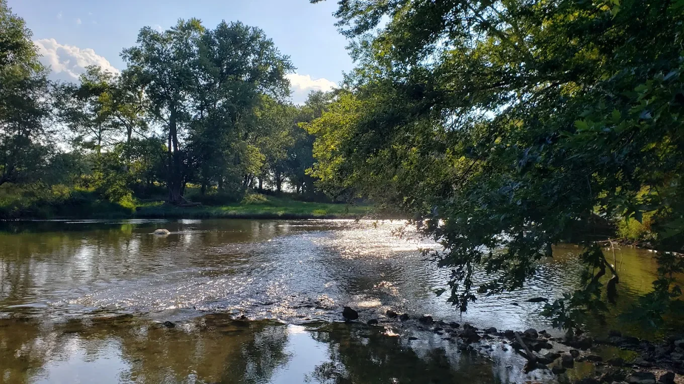 A peaceful river scene with trees on both sides, sunlight reflecting off the water, and a clear sky with clouds.