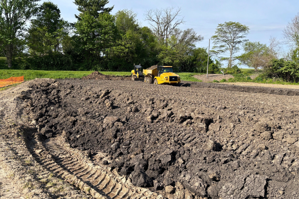 Construction site with a large pile of dirt and a yellow bulldozer, trees in the background, and clear sky.