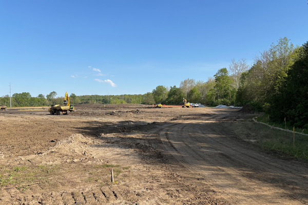 Construction site with earth-moving machinery on cleared land, trees on the right, clear blue sky.