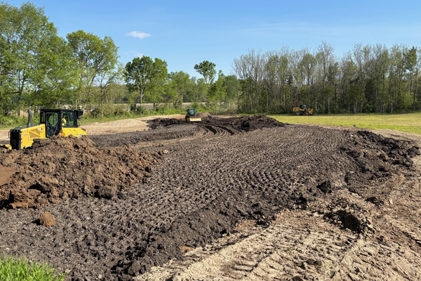 Construction site with bulldozers moving dirt in a grassy area surrounded by trees under a blue sky.