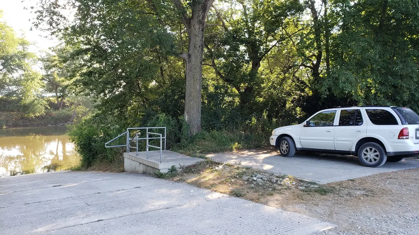 A white SUV parked near a small concrete ramp with metal handrails leading to a body of water, surrounded by tall green trees.