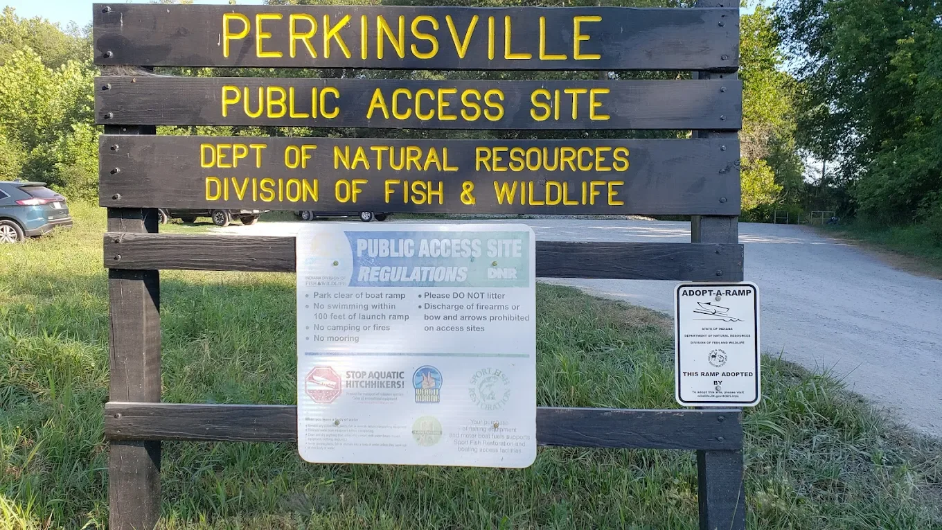 A large wooden sign at Perkinsville public access site with yellow lettering, indicating it is managed by the Indiana Department of Natural Resources, Division of Fish & Wildlife. Below it are smaller signs and notices about regulations, safety warni