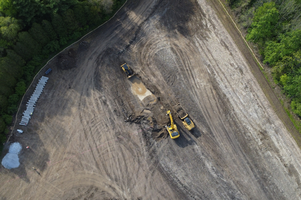 Construction site with three yellow excavators digging in dirt, surrounded by trees and tire tracks on the ground.