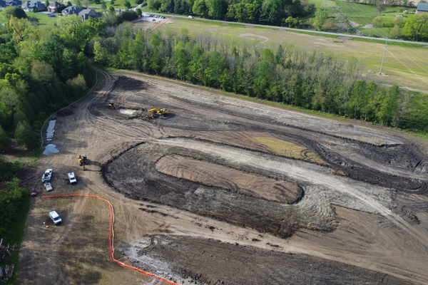 Aerial view of a construction site with earth-moving machinery creating a racetrack-shaped excavation amidst cleared land and surrounding trees.