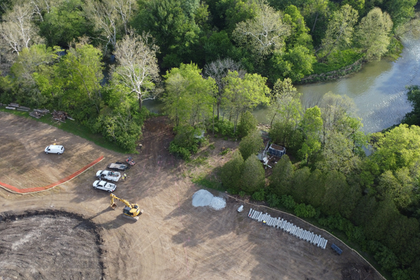 Construction site near a wooded area and a river, with parked cars, a small excavator, and materials stacked on the ground.