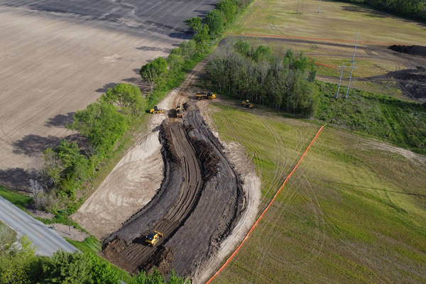Aerial view of a construction site with excavators working on a curved dirt road, surrounded by green fields and trees, with power lines in the distance.