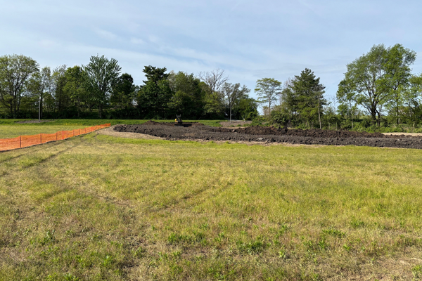 A grassy field with patchy green grass, a section of dark tilled soil, and orange fencing along the left side, with trees and a cloudy sky in the background.