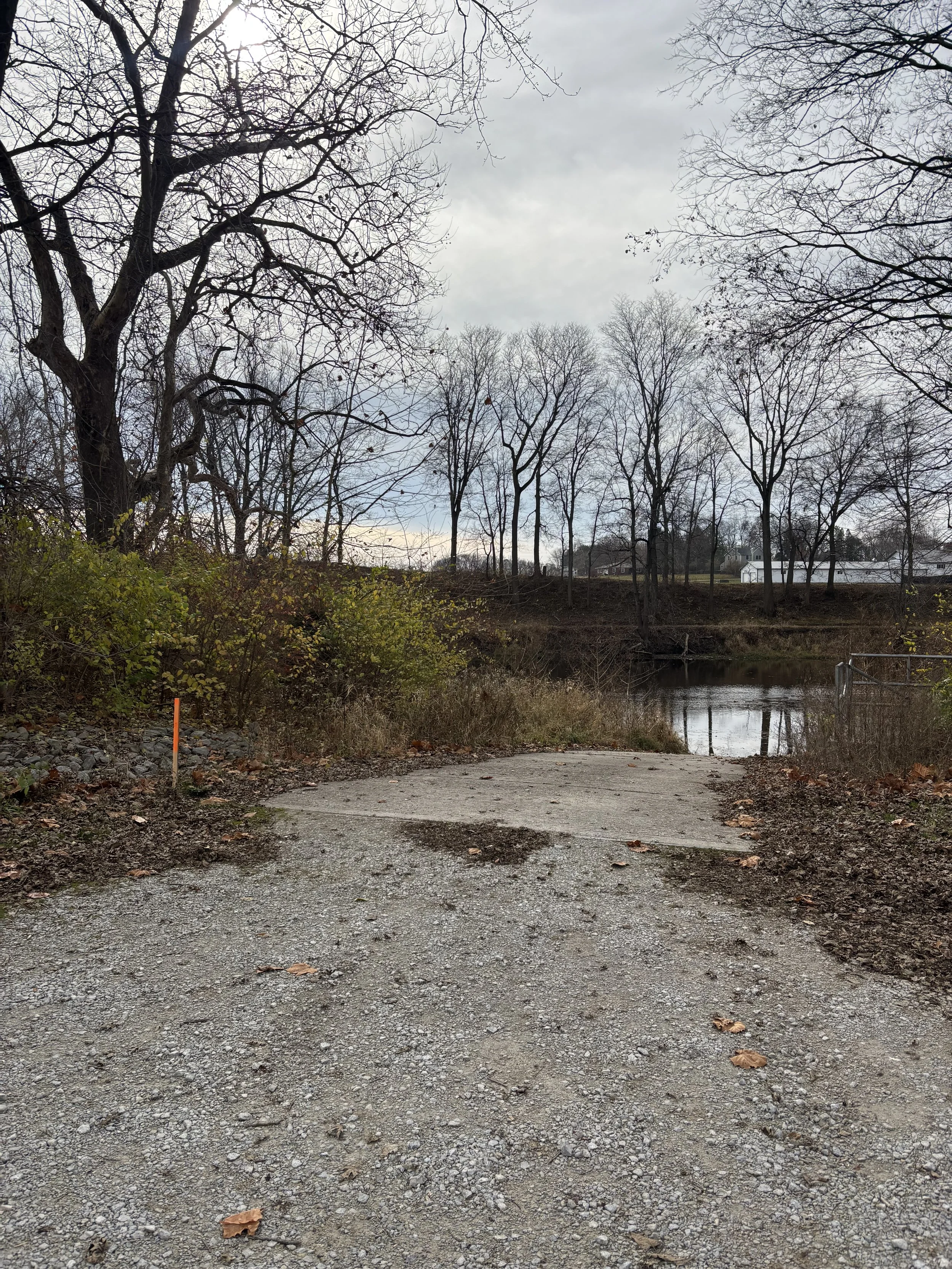 A gravel pathway leading to a body of water, with leafless trees on either side and overcast sky overhead.