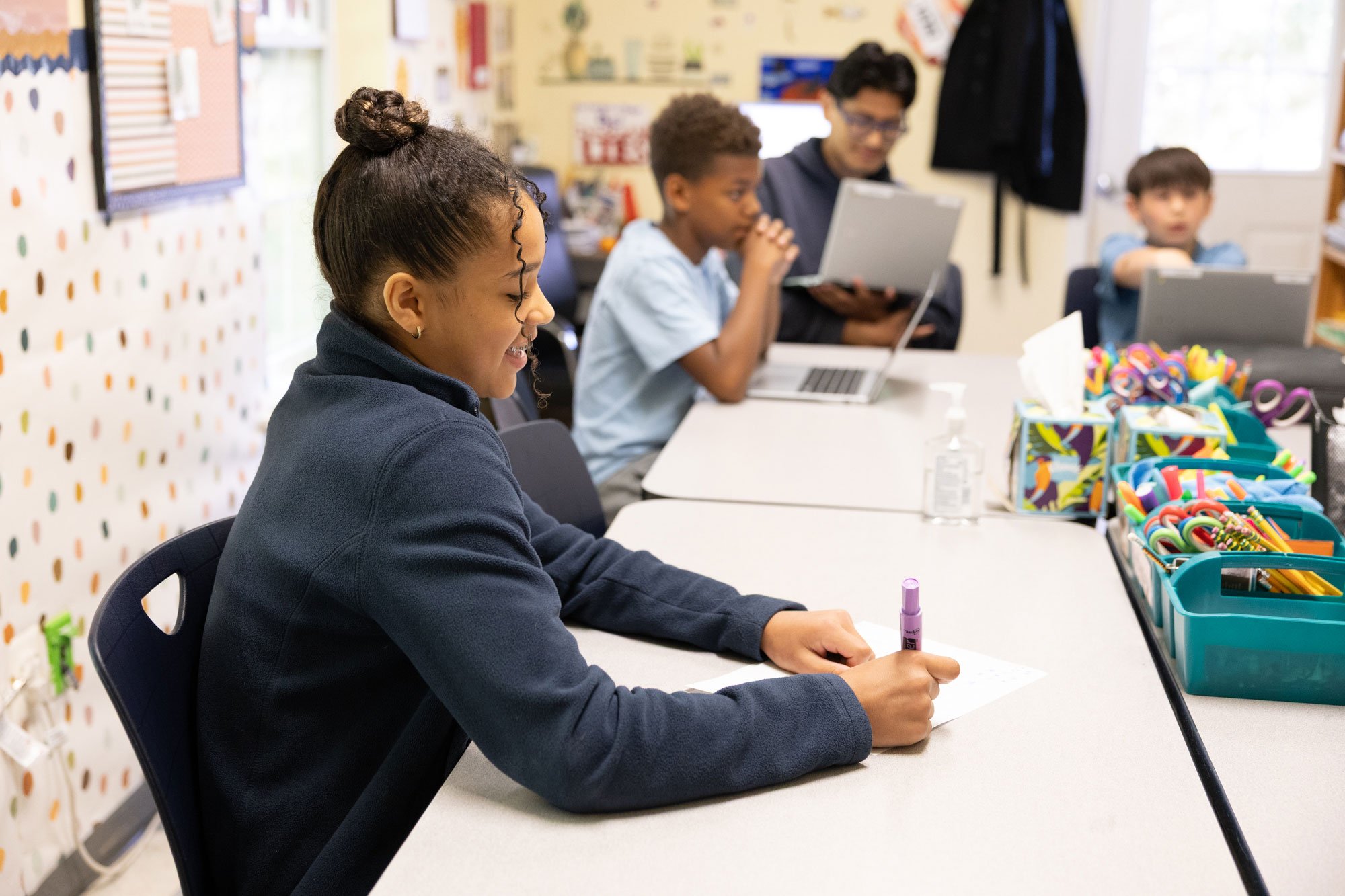 A group of students in a classroom, sitting at a table with art supplies, writing and using laptops, smiling and focused on their activities.