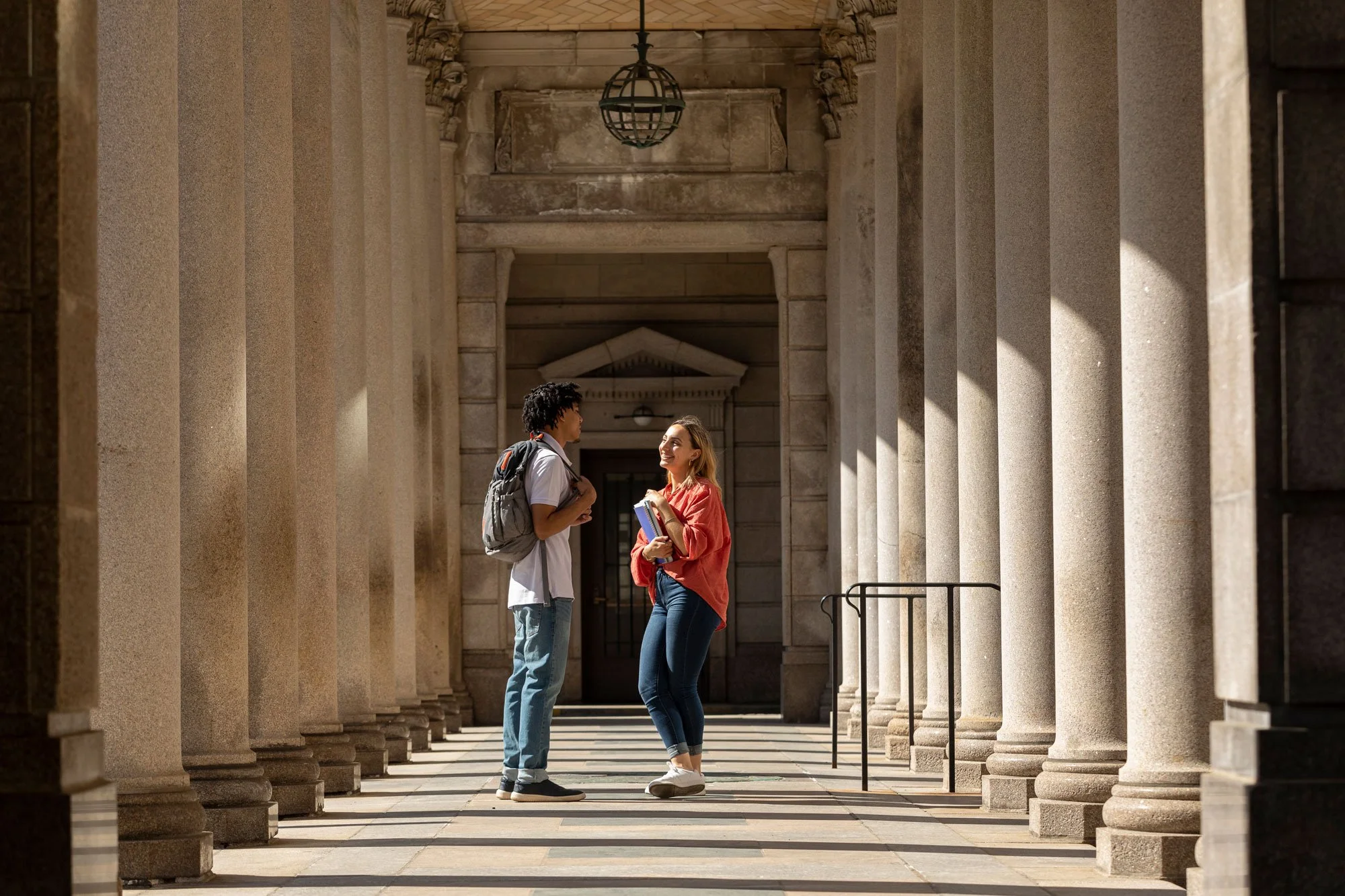 A man and woman are standing face-to-face in an outdoor corridor with tall stone columns. The man, wearing a white shirt and backpack, is listening, while the woman, in a red jacket and jeans, is smiling and holding a notebook.
