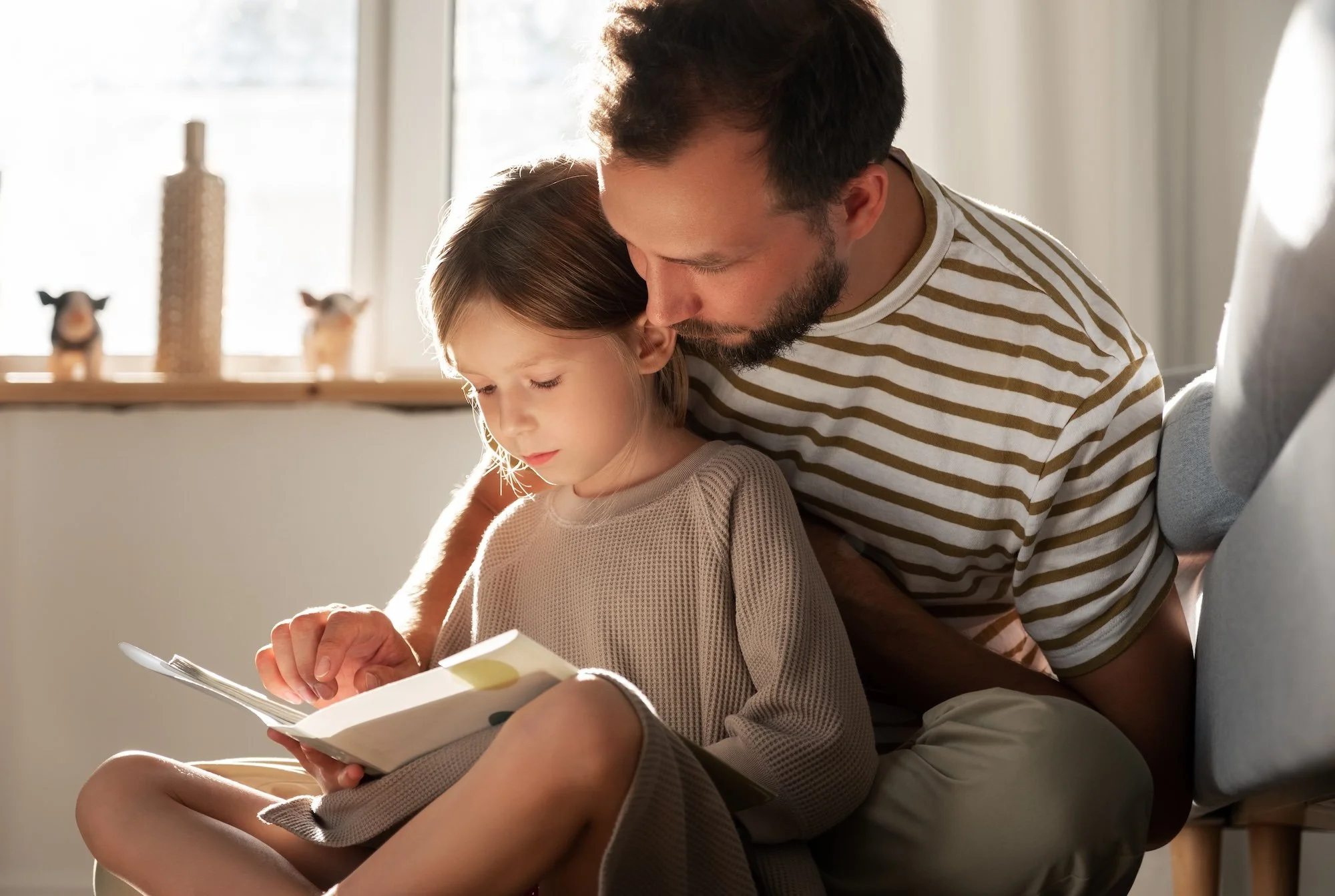 A man and a young girl sitting closely on a couch, with the man leaning his head against the girl's and looking at a book she is holding.