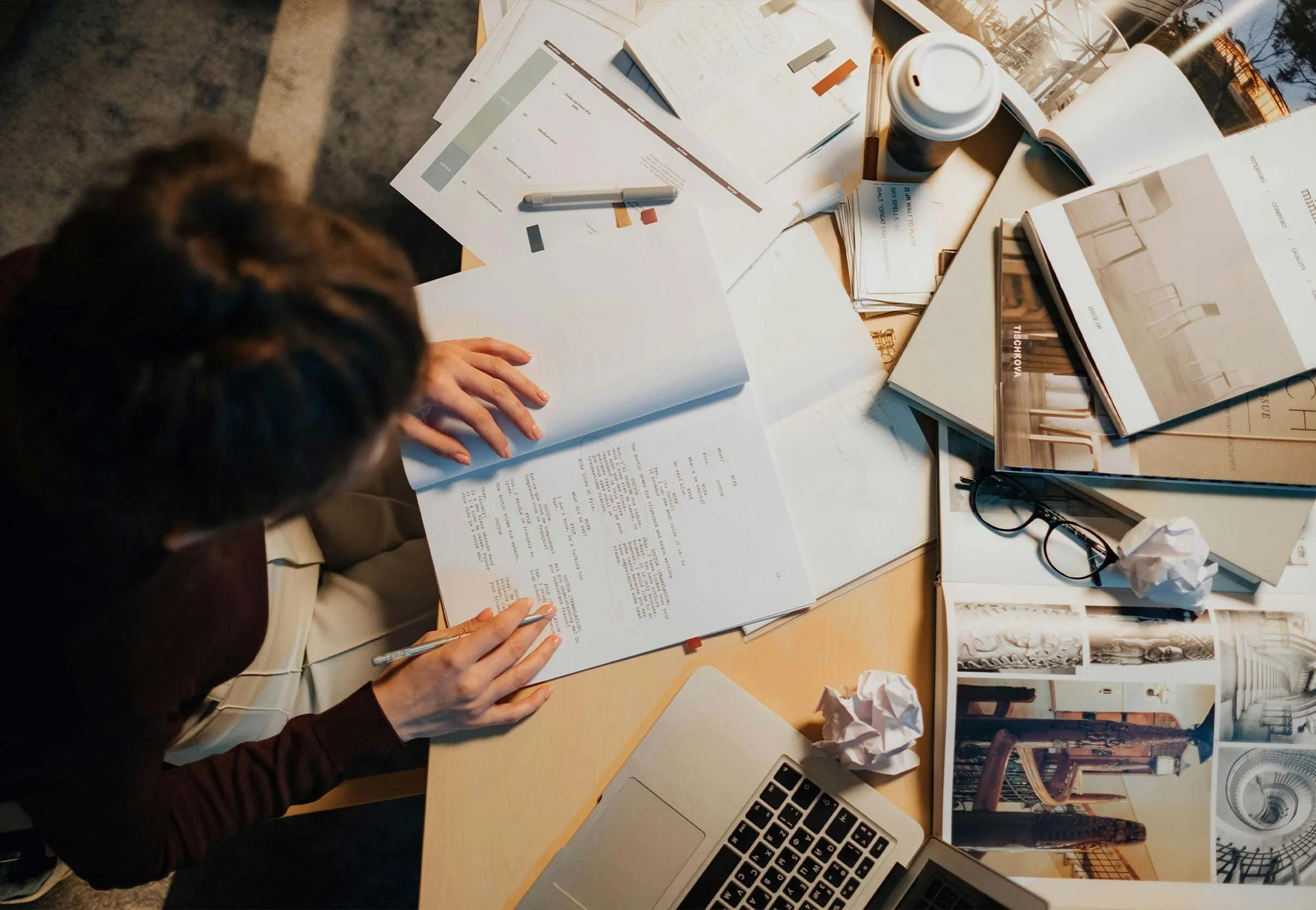 Overhead view of a person working at a cluttered desk with papers, books, magazines, crumpled paper, a cup of coffee, eyeglasses, a pen, and a laptop.