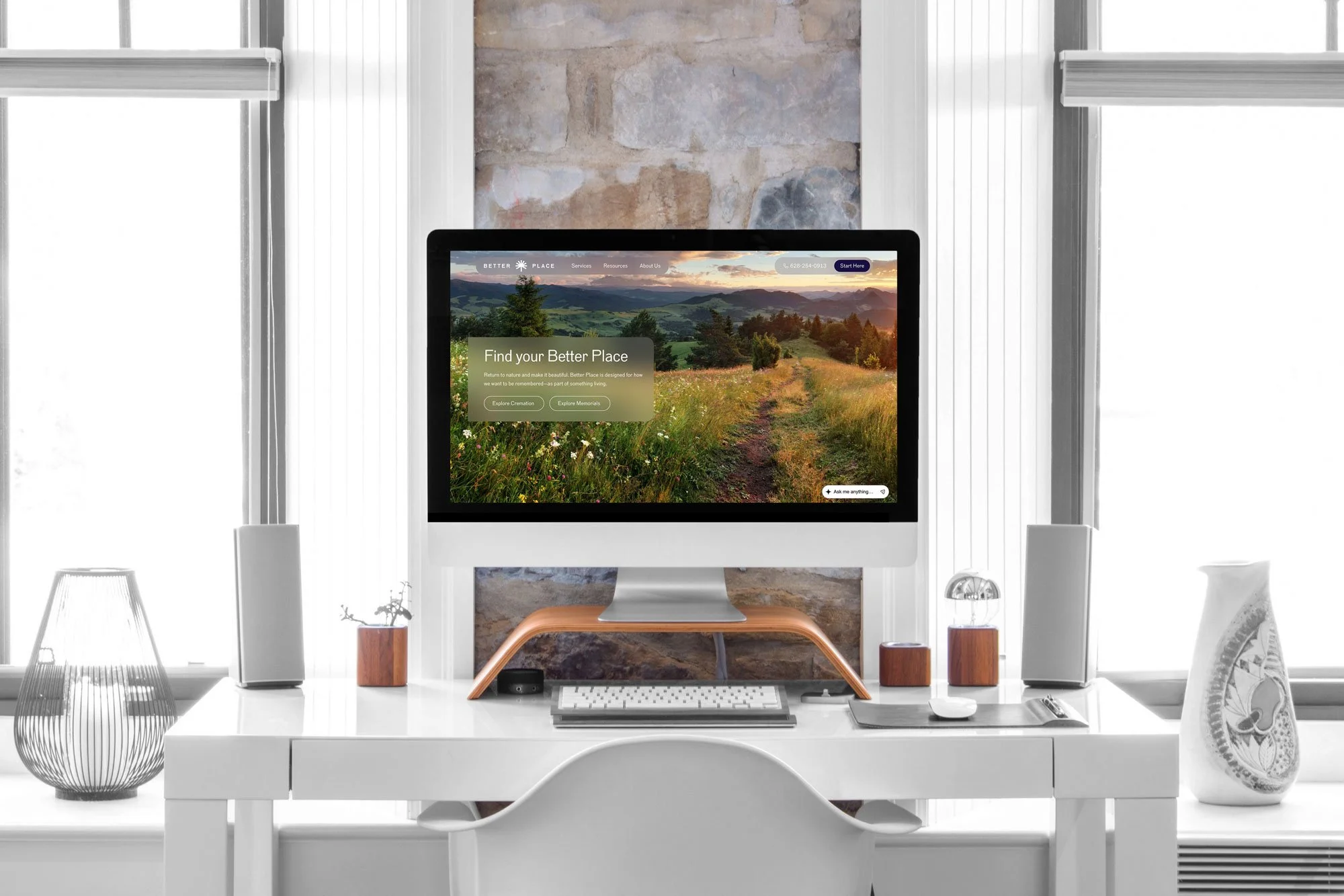 A modern workspace with a white desk, computer monitor displaying a landscape of green hills and a sunset, and decorative objects including a wire vase, a wooden plant pot, and a decorative object on a side table. The background features a stone wall and large windows with white blinds.