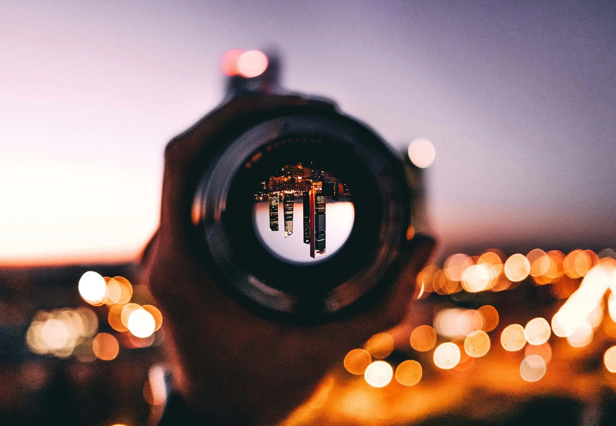 A camera lens aimed at the city skyline during dusk, capturing the illuminated buildings and city lights, with a blurred background and bokeh effect.