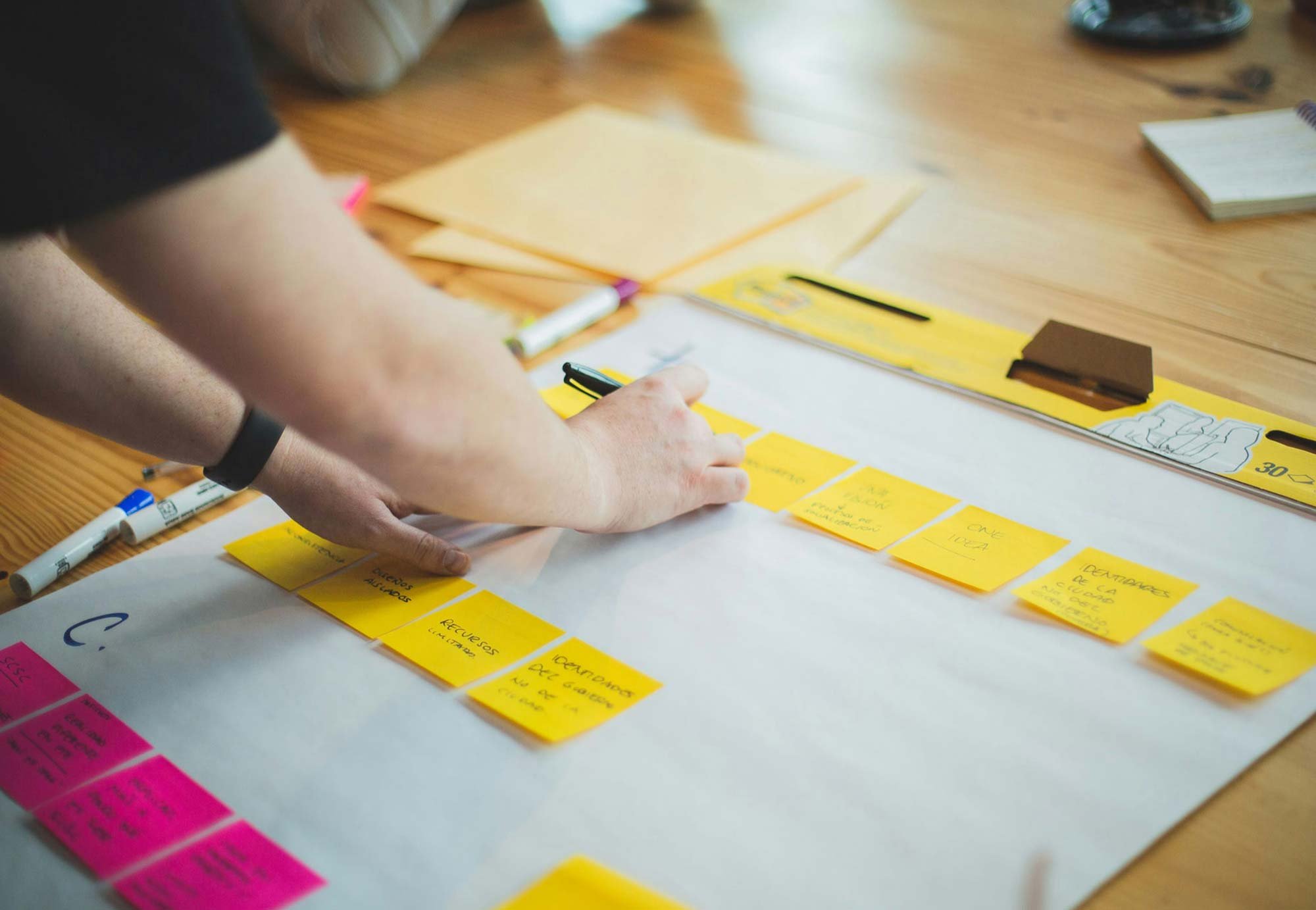 A person arranging yellow sticky notes on a large white sheet on a wooden table, with various markers, notebooks, and paper stacks nearby.