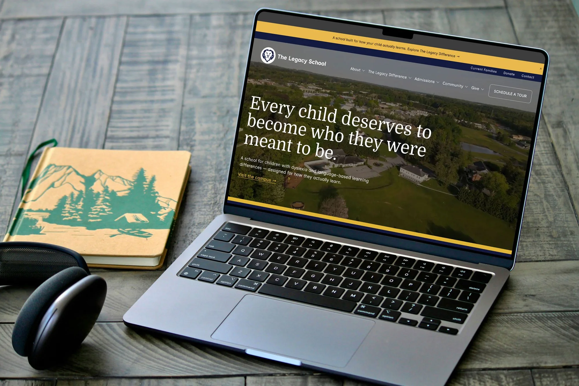 Laptop displaying a website for The Legacy School, with a background of green trees and buildings, on a wooden desk with a notebook, headphones, and a speaker nearby.